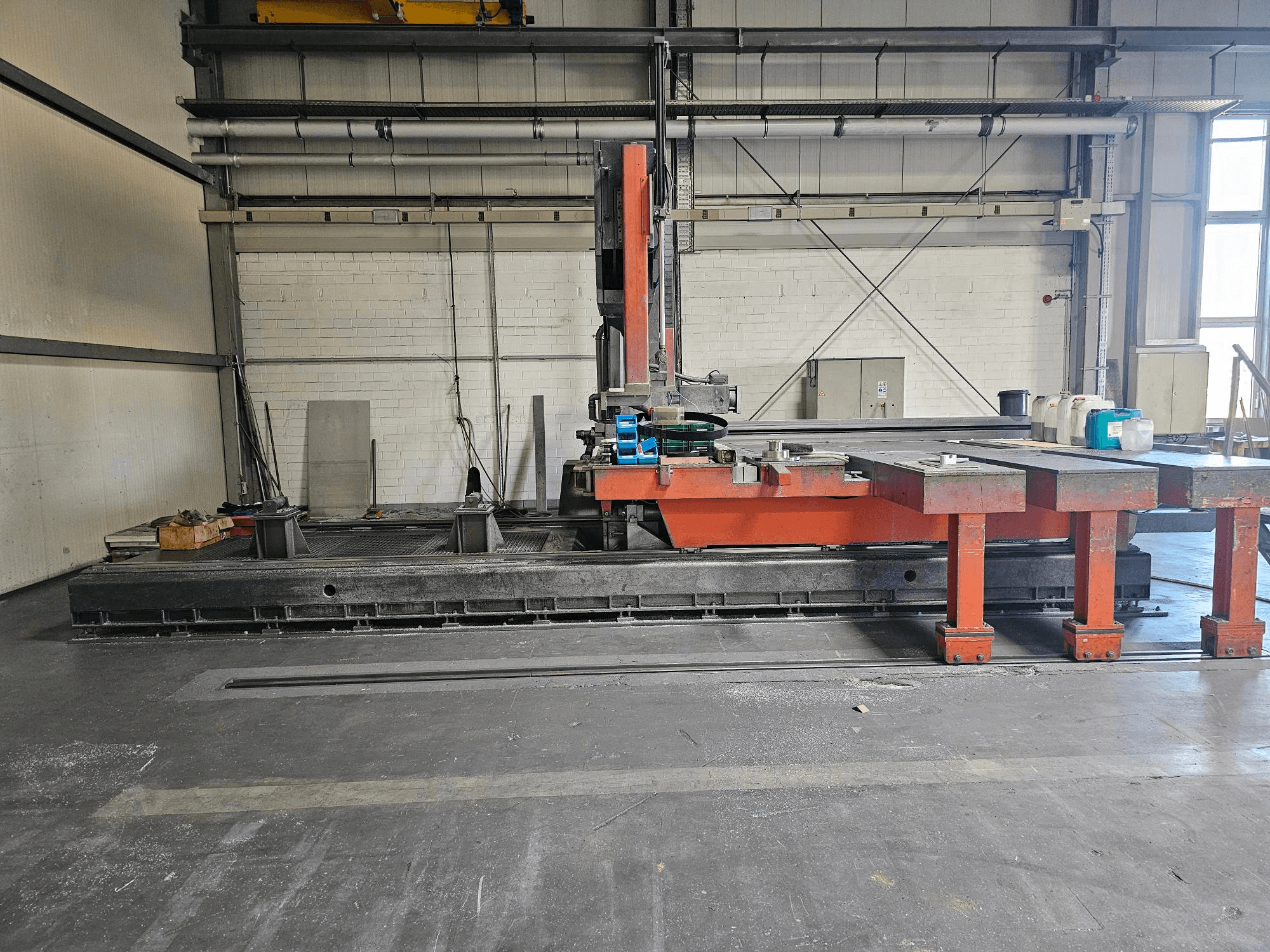 Przemysłowa maszyna do cięcia w otwartym warsztacie, pomarańczowo-czarna z dużym stołem; widok z boku pokazujący akcesoria i przestrzeń roboczą.