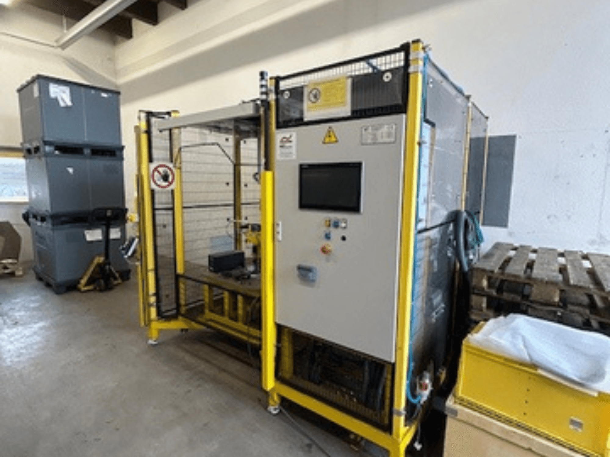 Maszyna przemysłowa z panelem sterowania i zabezpieczeniami, widok z przodu; żółta rama, szare pojemniki i palety w tle.