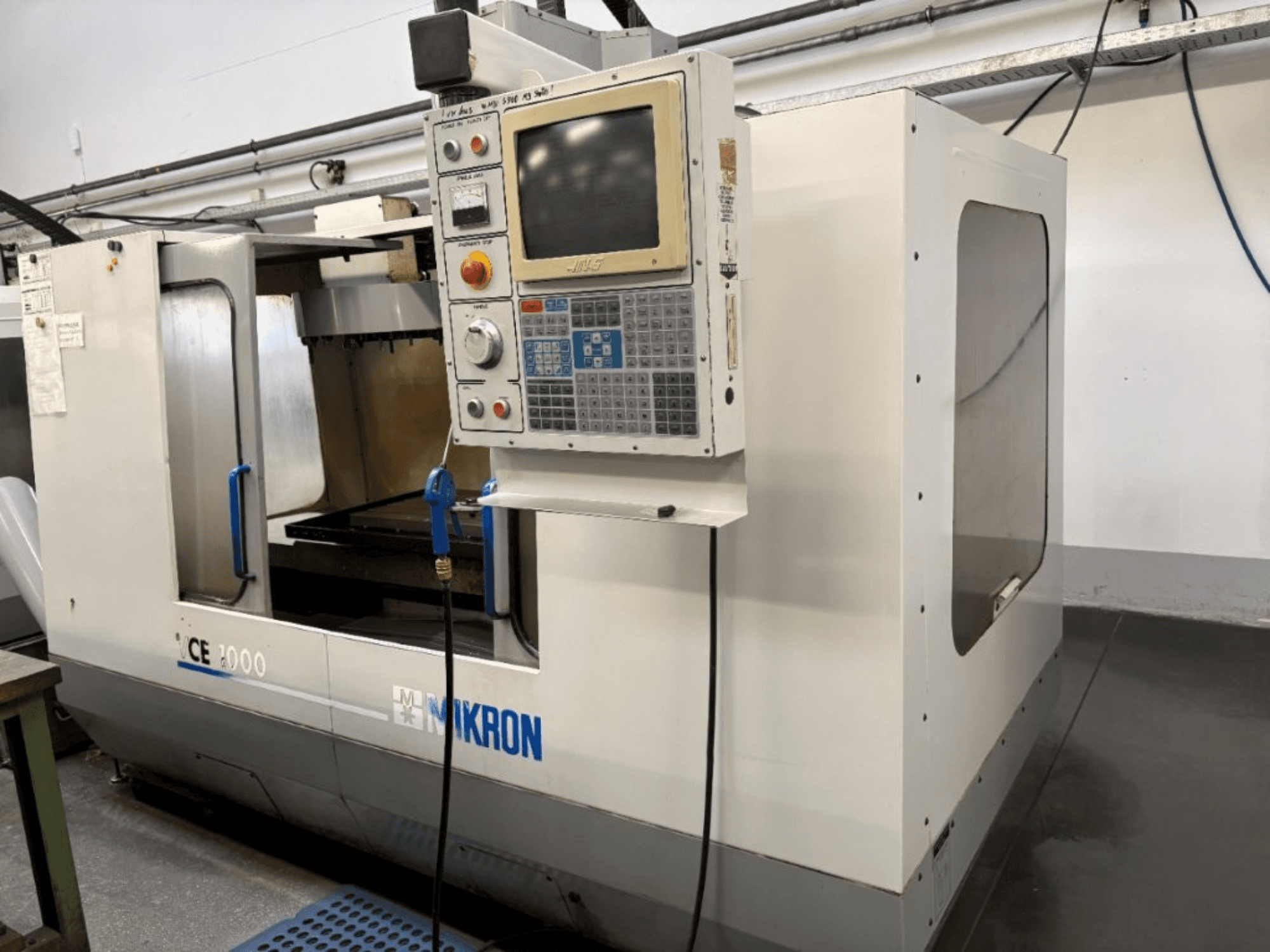Maszyna CNC Mikron CE 2000, widok z przodu; posiada panel sterowania z przyciskami i ekranem, srebrną obudowę i obszar roboczy.