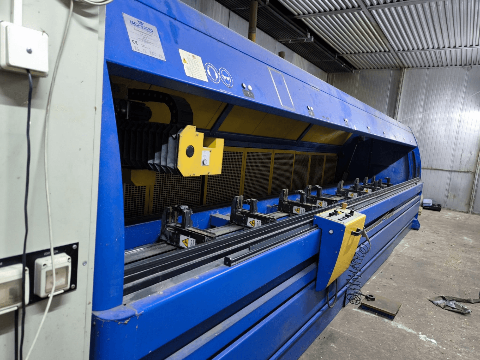 Przemysłowa przecinarka Schneider w kolorze niebieskim, widok z boku, z widocznym panelem sterowania, pasami i wieloma stacjami zaciskowymi.