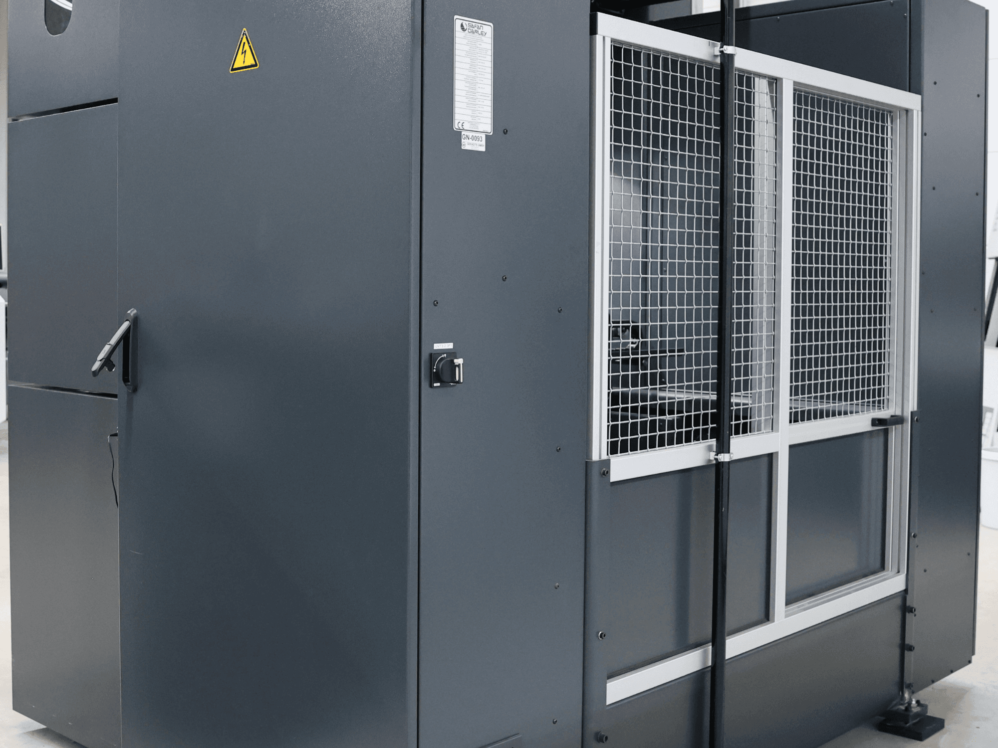 Szara maszyna przemysłowa z ostrzeżeniem o bezpieczeństwie, widok z przodu pokazujący siatkowe drzwi i panel sterowania, model oznaczony etykietą.