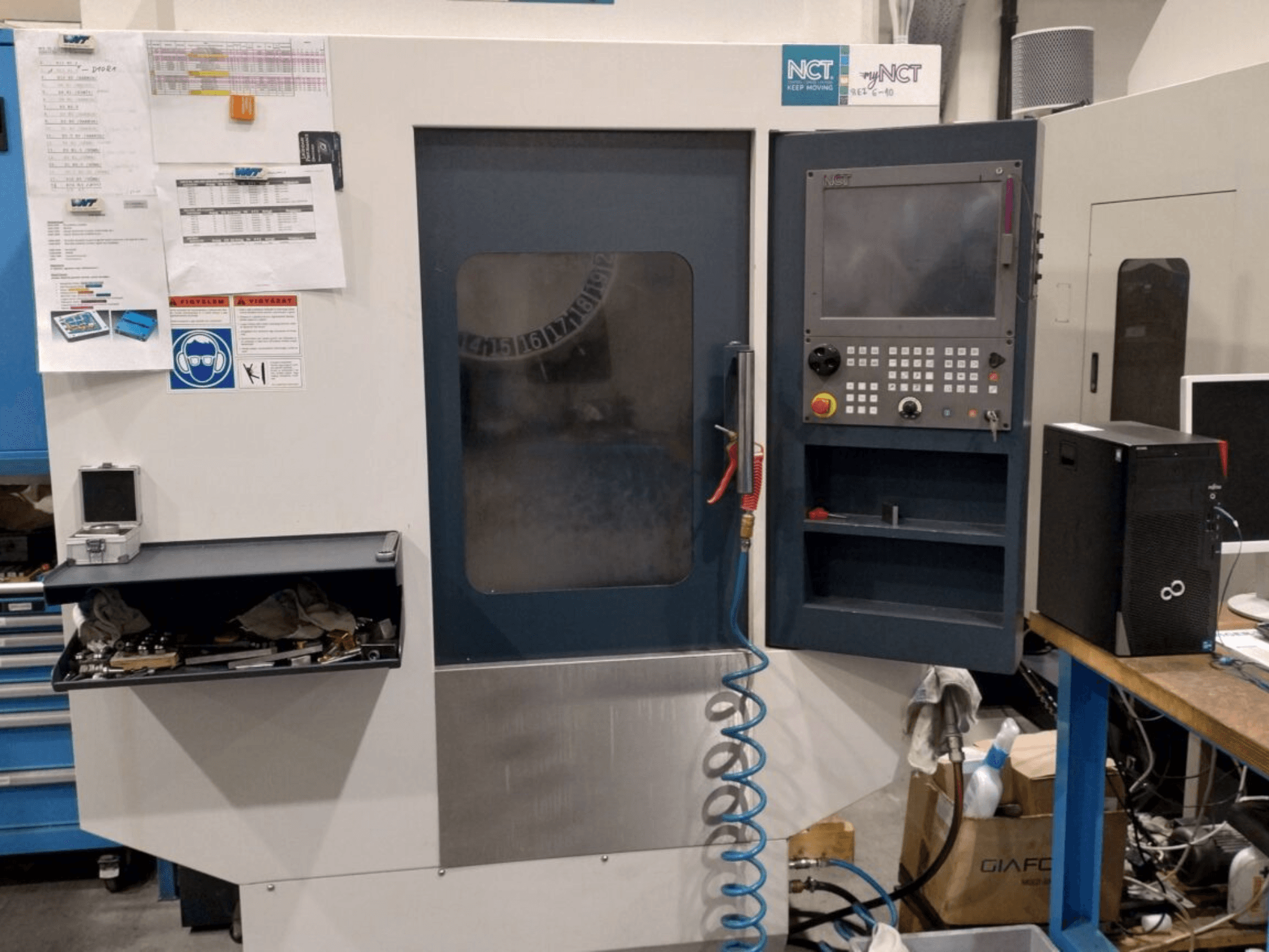 Maszyna NCT CNC, widok z przodu, panel sterowania z klawiaturą numeryczną, narzędzia w szufladzie i instrukcja obsługi na ścianie.