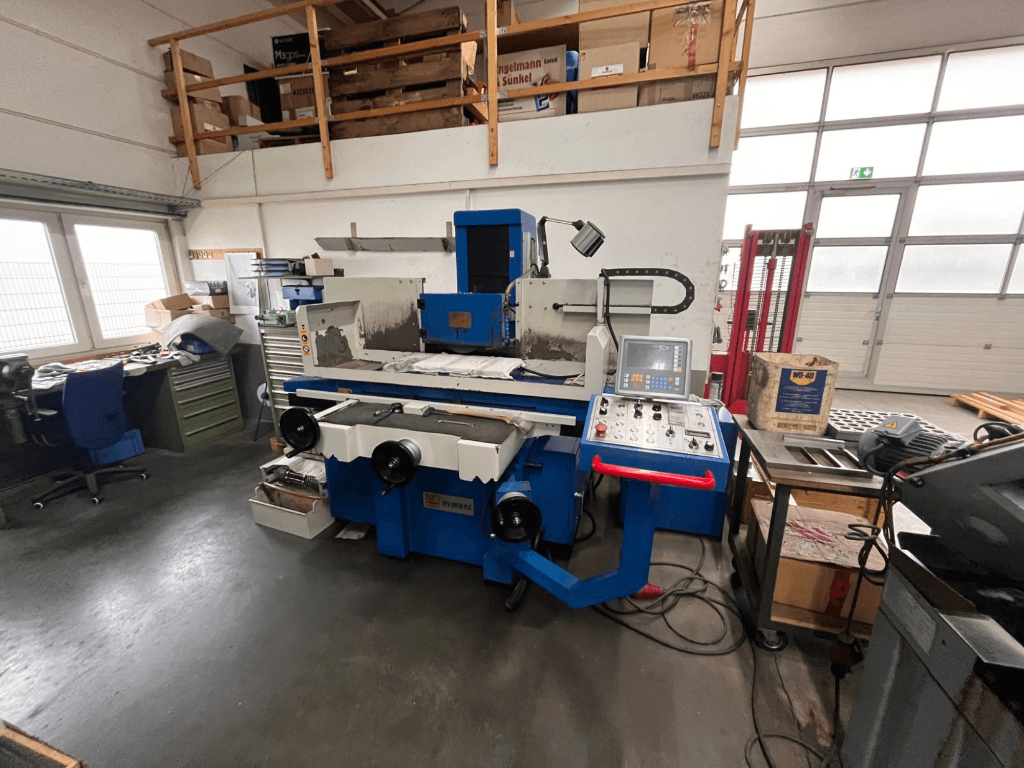 Niebieska szlifierka do płaszczyzn z cyfrowym panelem sterowania, widok z przodu, pokazujący bałagan w przestrzeni roboczej i zieloną szafkę narzędziową.