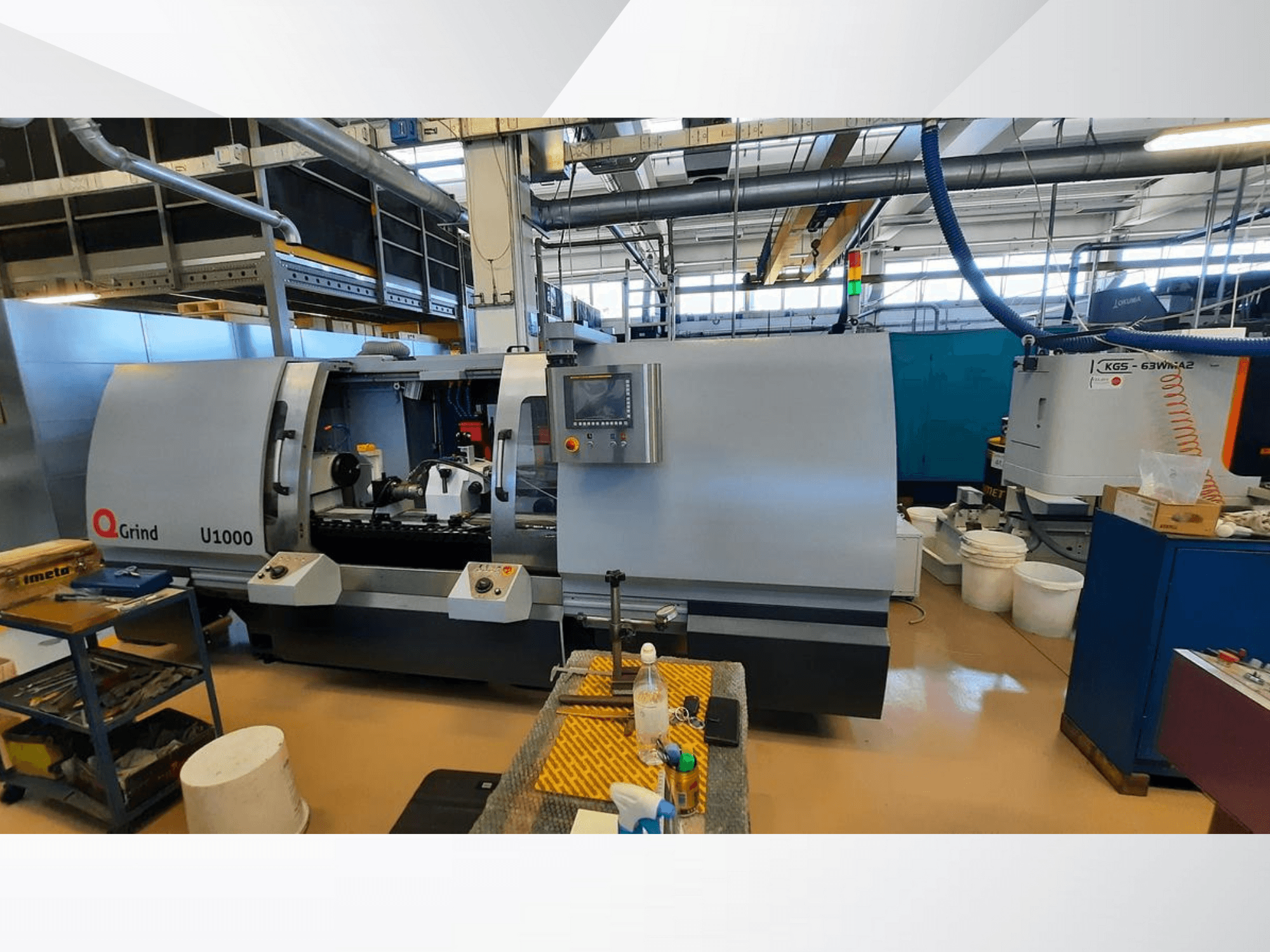 Maszyna Q-Grind U1000 w warsztacie, pokazująca widok z przodu z panelem sterowania i otwartym obszarem cięcia, otoczonym sprzętem.