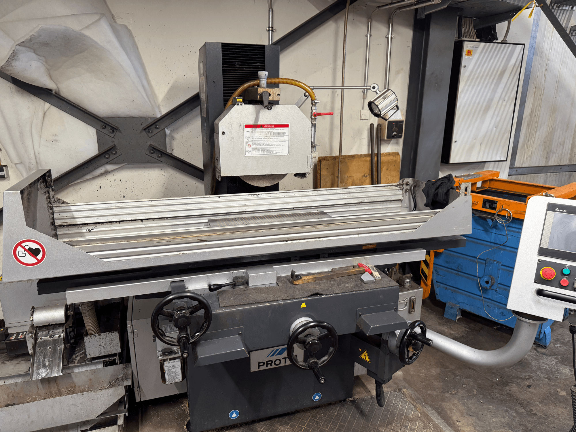 Maszyna PROTEC, widok z przodu, pokazująca szlifierkę do płaszczyzn z ręcznymi elementami sterującymi, ostrzeżeniami dotyczącymi bezpieczeństwa i układem chłodzenia.