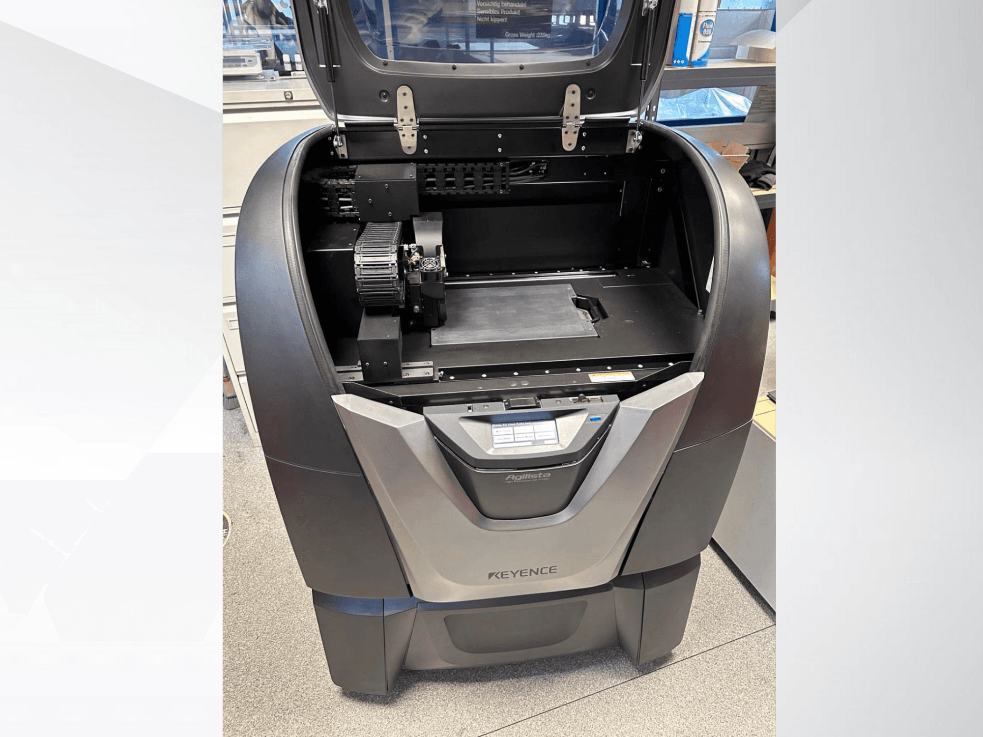 Drukarka 3D KEYENCE Agilista, widok z przodu, ujawnia otwartą górę, dużą platformę roboczą i wewnętrzne komponenty mechaniczne.