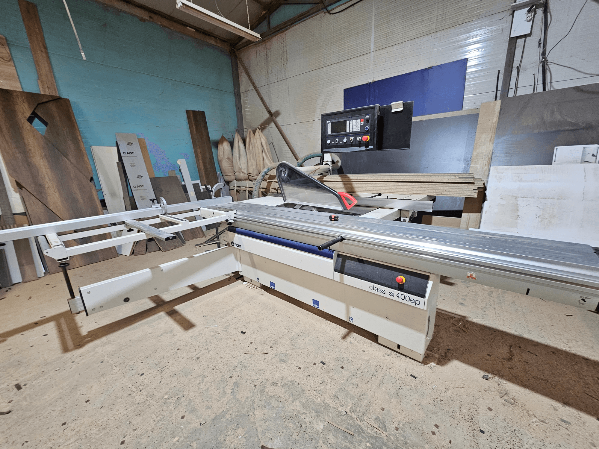 Piła panelowa klasy SI400EP, widok z przodu, z regulowaną przykładnicą, ustawiona na piaszczystej podłodze warsztatu, otoczona drewnianymi panelami.