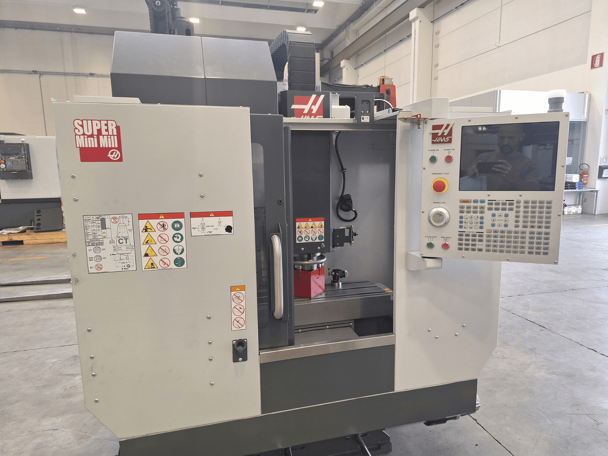 Widok z przodu frezarki HAAS Super Mini Mill przedstawiający panel sterowania, etykiety bezpieczeństwa oraz obszar obróbki z zamontowanym elementem obrabianym.