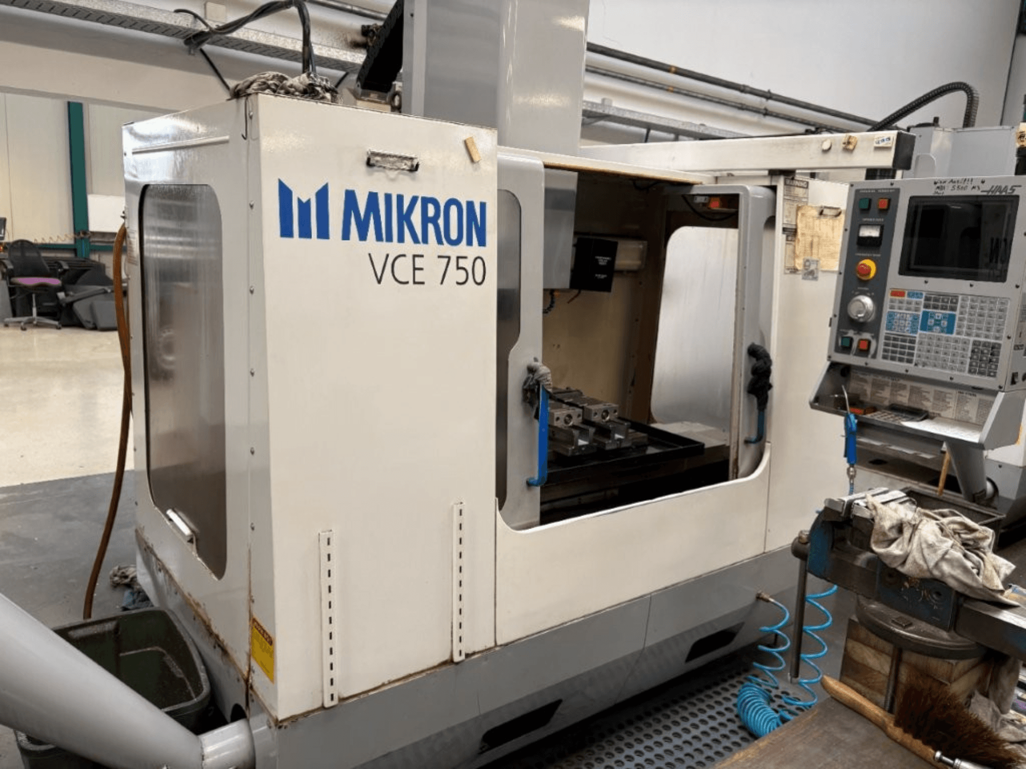 Frezarka CNC Mikron VCE 750 - widok z przodu, z obrobionym stołem roboczym i panelem sterowania z różnymi przyciskami i ekranami.