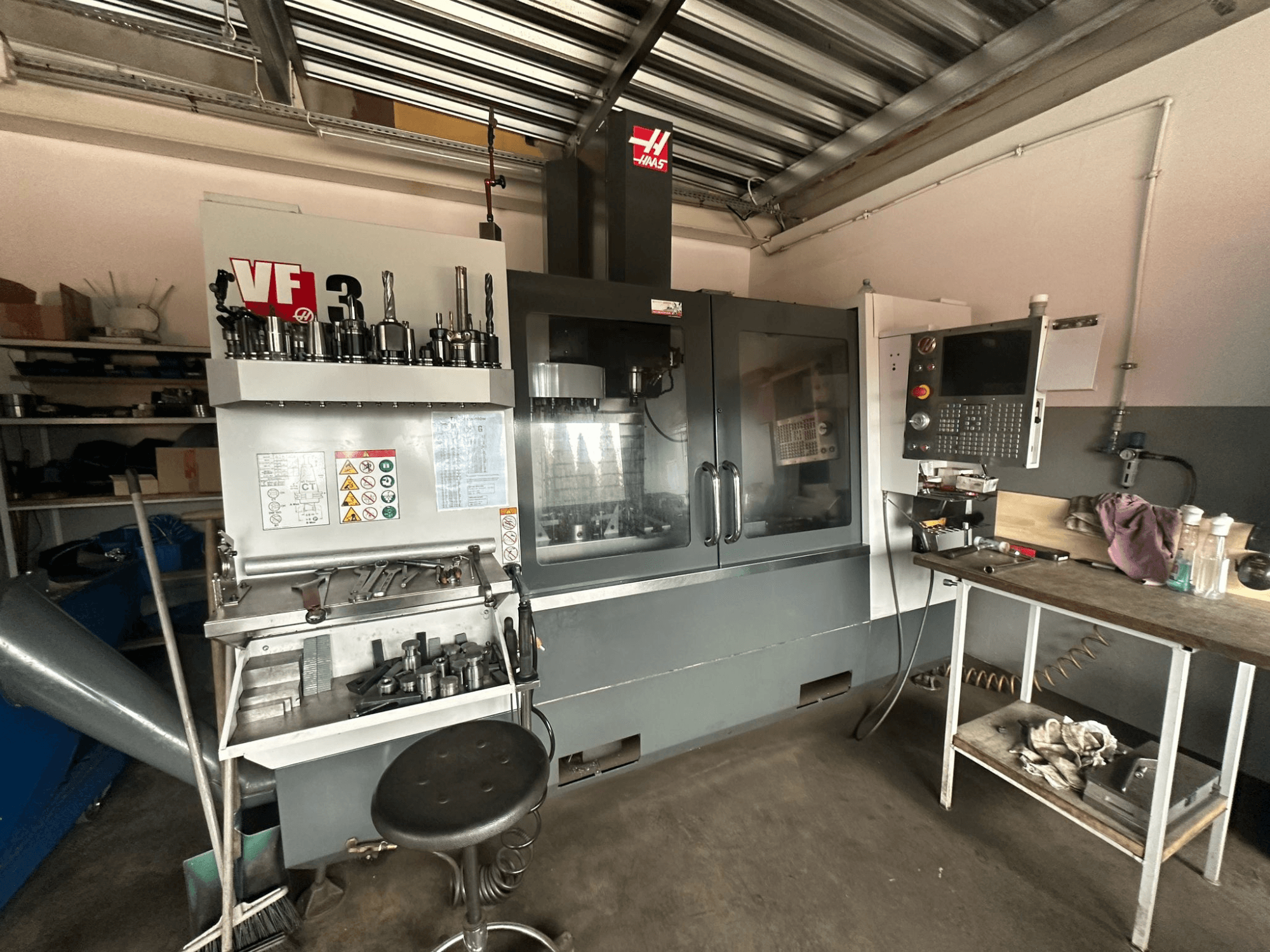 Maszyna CNC Haas VF-3 w warsztacie, widok z przodu z narzędziami i elementami sterującymi na sąsiednim stole, wyświetlanie interfejsu technicznego i informacji dotyczących bezpieczeństwa.