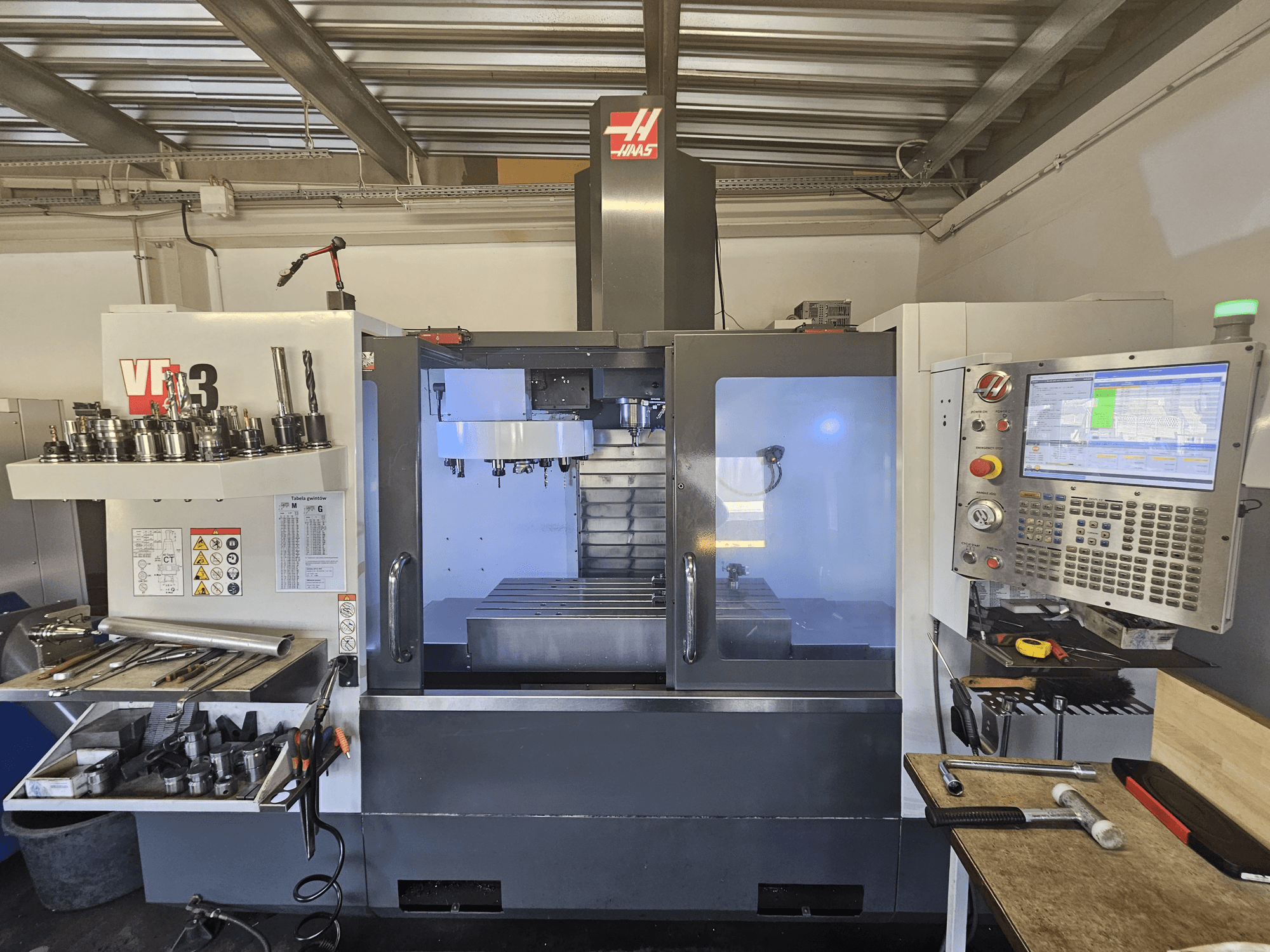 Maszyna CNC Haas VF-3 w warsztacie, widok z przodu przedstawiający narzędzia, elementy sterujące i obszar frezowania z oświetlonym wnętrzem.