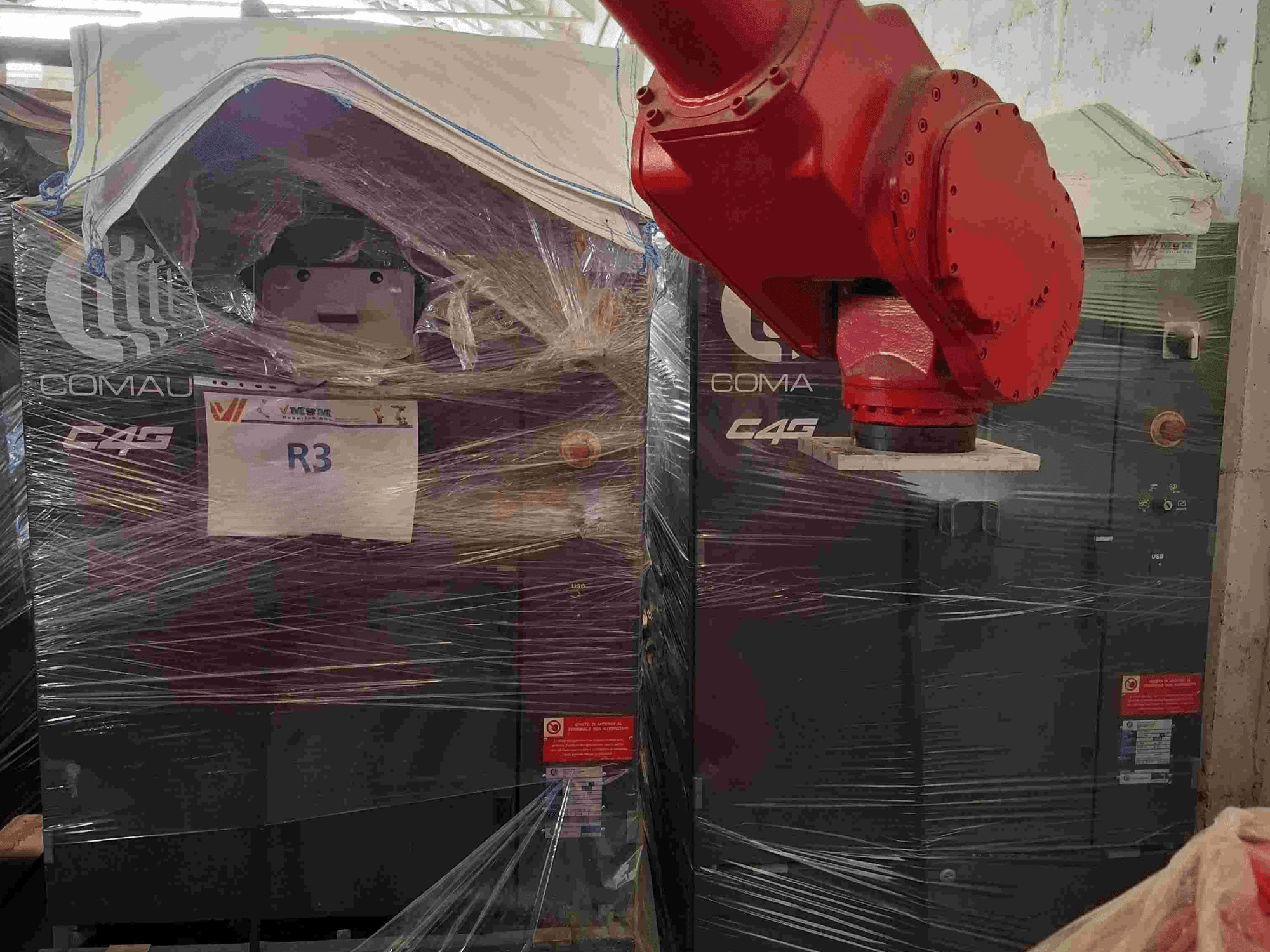 Ramię robotyczne Comau C4G widziane od przodu, owinięte w plastik, oznaczone jako "R3", z naklejkami ostrzegawczymi widocznymi na maszynie.