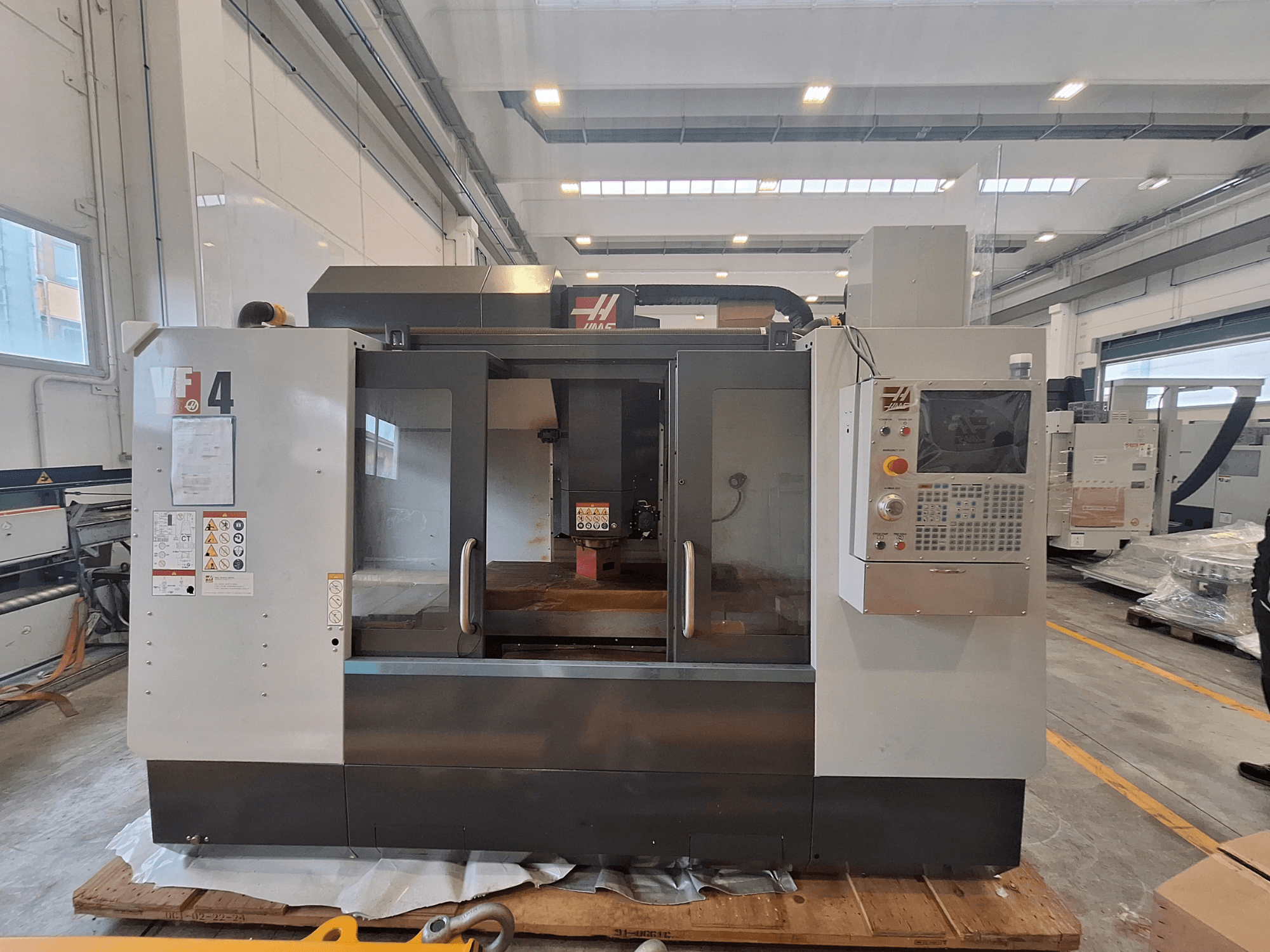 Pionowe centrum obróbcze HAAS VF-4, widok z przodu, przedstawiający panel sterowania, obszar roboczy oraz etykiety bezpieczeństwa w otoczeniu magazynowym.