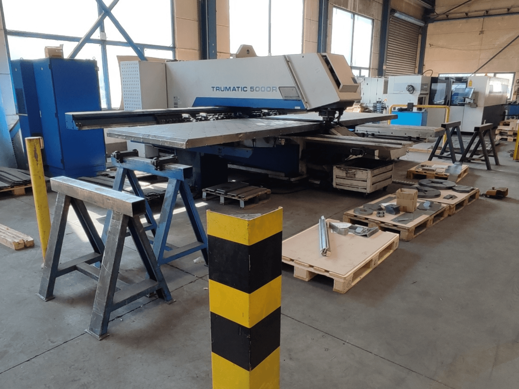 Wykrawarka CNC TRUMPF Trumatic 5000 R w warsztacie, widok z boku, z narzędziami i materiałami na paletach w pobliżu.