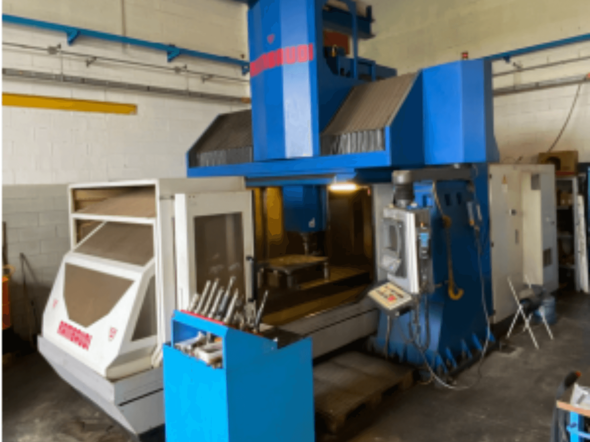 Pionowe centrum obróbcze Rambaudi RF103, widok z przodu pokazujący elementy sterujące, magazyn narzędzi i obszar obróbki. Niebiesko-biała obudowa.