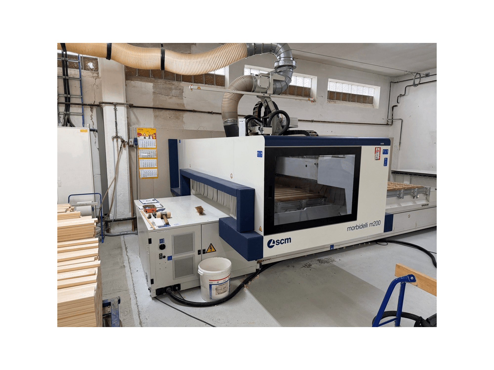 Centrum obróbcze CNC SCM Morbidelli M200F w warsztacie, widok z boku z kawałkami drewna na stole i systemem wentylacyjnym nad nim.