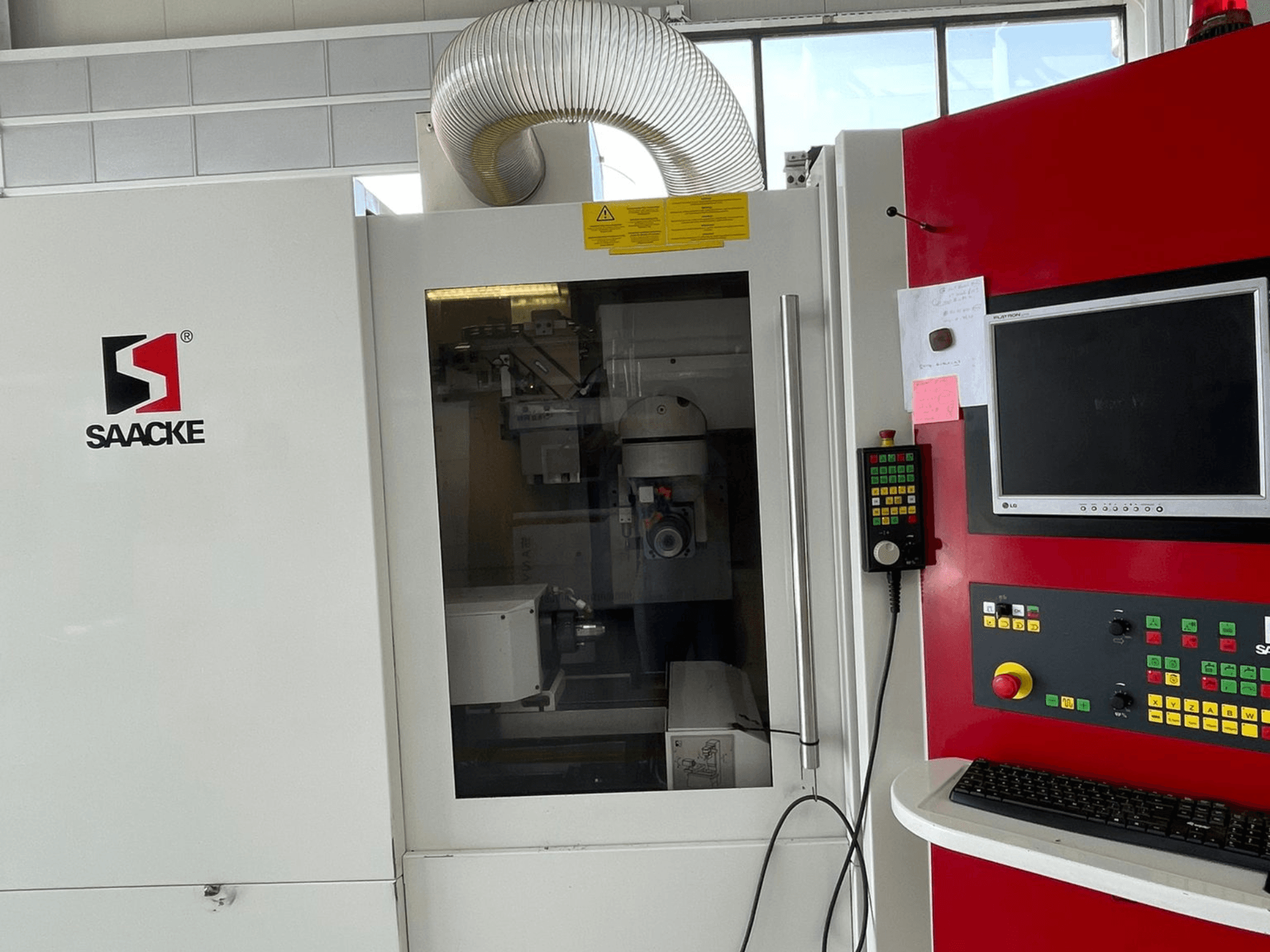 Maszyna SAACKE w kolorze białym i czerwonym, widok z przodu, z panelem sterowania, monitorem i widocznymi przez szybę częściami wewnętrznymi.