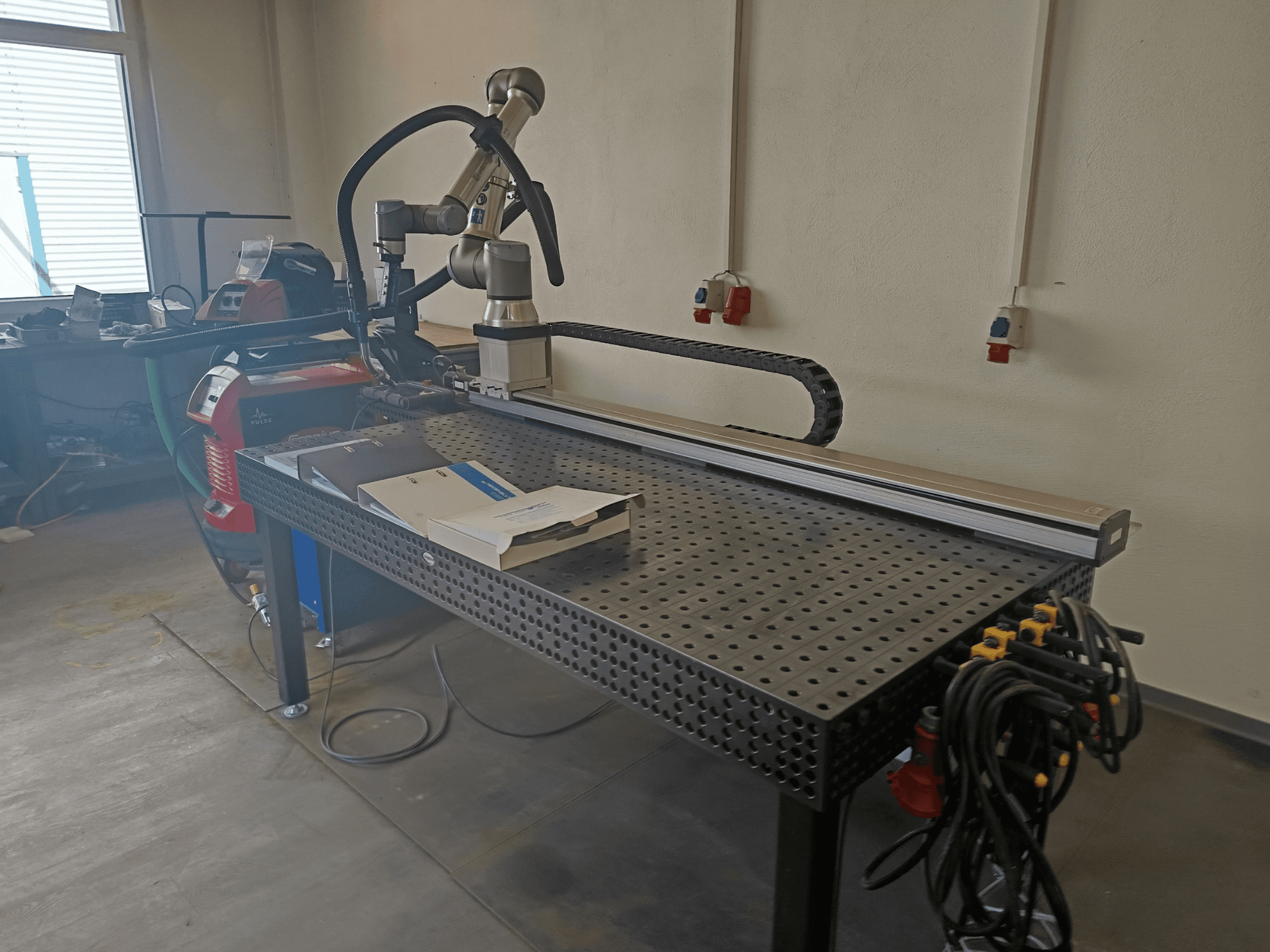 Komora spawalnicza FRONIUS TransSteel 4000 do spawania impulsowego widziana z boku, wyposażona w ramię robota, obszar roboczy oraz przyłącza zasilające.