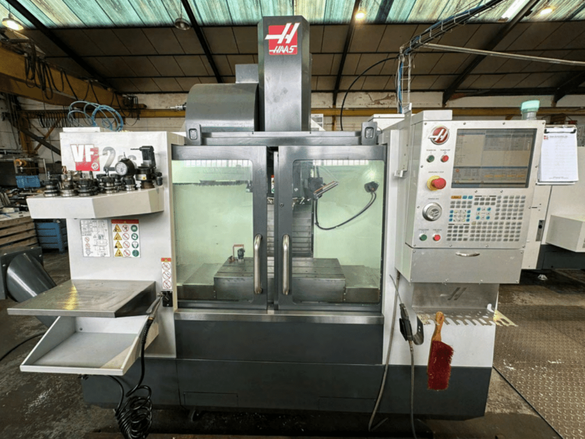 Widok maszyny CNC Haas VF2 z przodu, pokazujący panel sterowania, uchwyty narzędzi i obszar roboczy z etykietami bezpieczeństwa i szczotką.