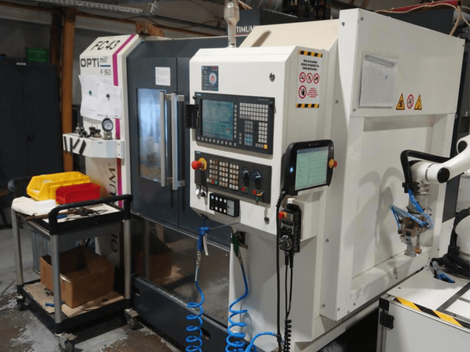 Panel sterowania frezarki OPTIMUM F50 z wężami, narzędziami i ramieniem robota widocznym w warunkach warsztatowych.