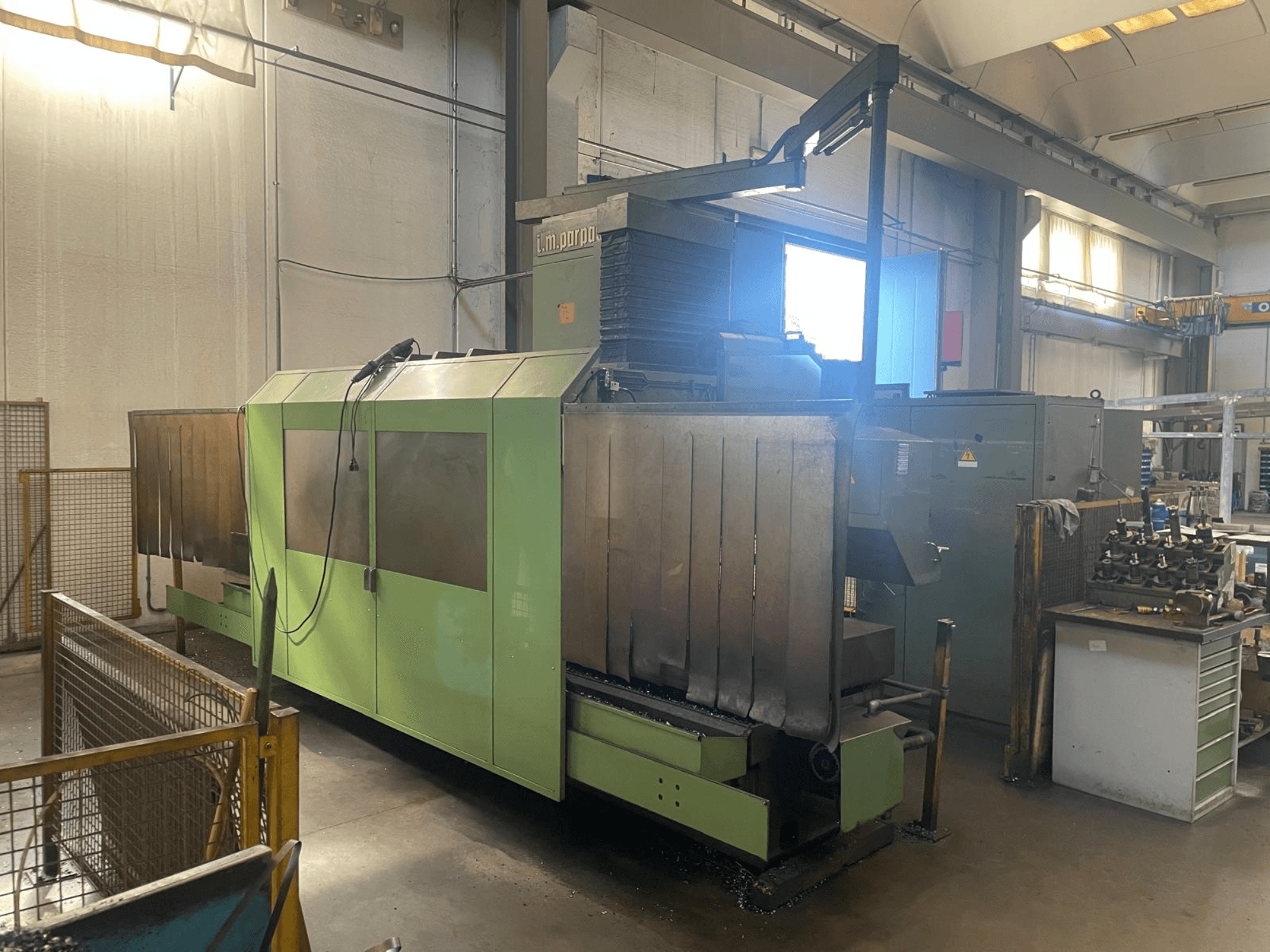 Zielona maszyna przemysłowa z widocznymi metalowymi komponentami, widok z przodu; posiada panel sterowania, ostrzeżenia bezpieczeństwa i stację roboczą w pobliżu.