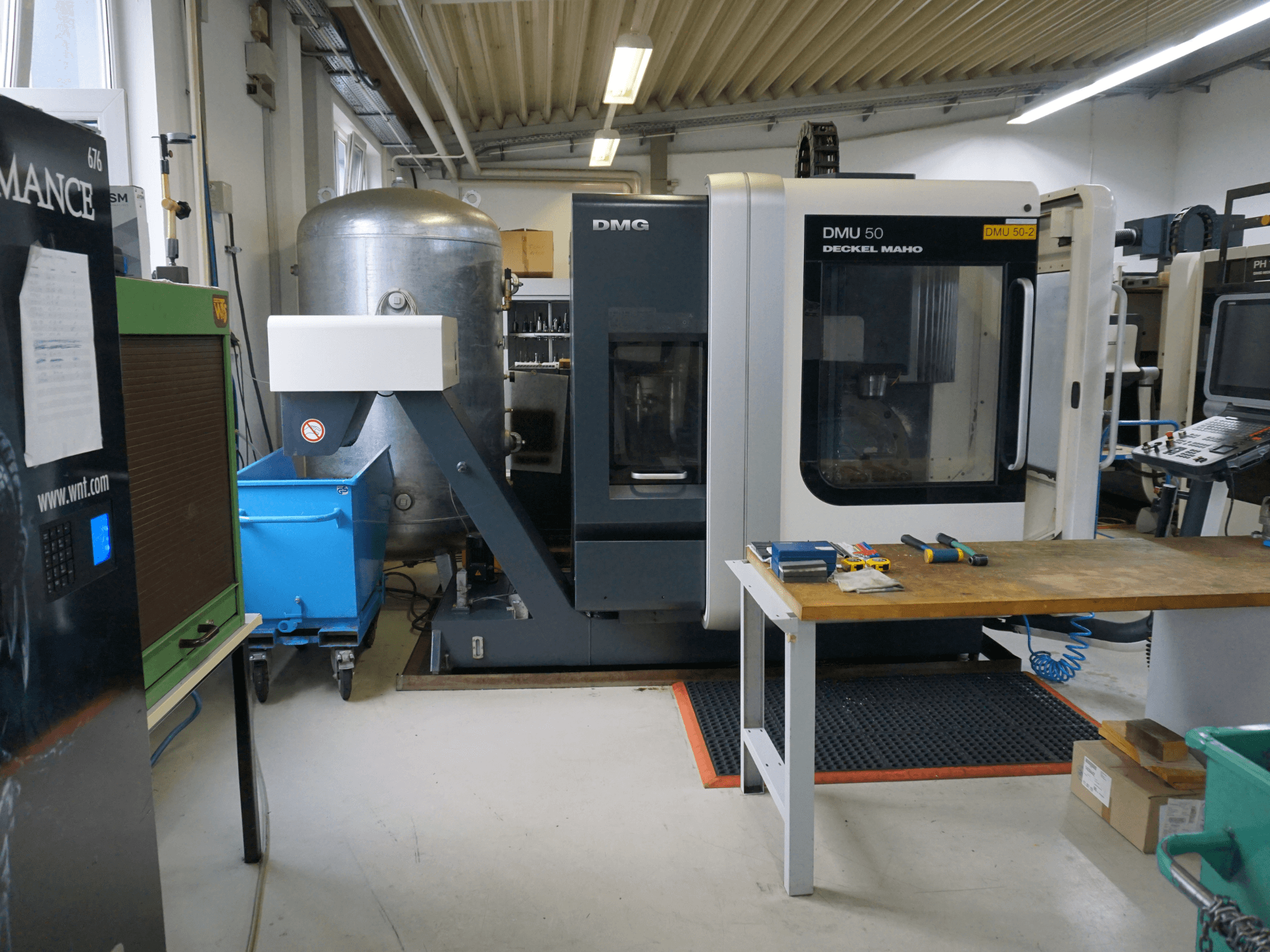 Frezarka DMG DMU 50 w warsztacie, widok z przodu, z narzędziami na drewnianym stole i niebieskim pojemnikiem na kółkach w pobliżu.
