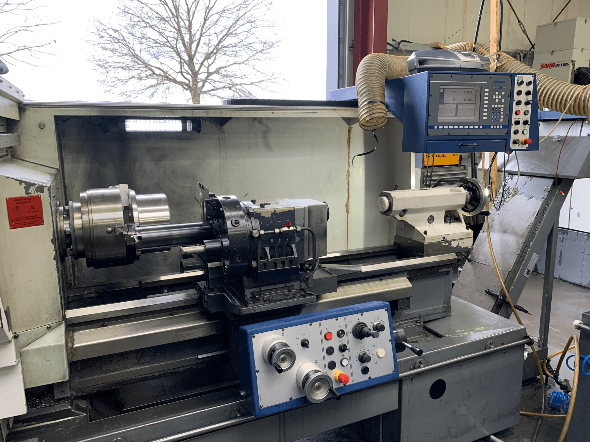 Tokarka pozioma Weiler E55X1000 w warsztacie, pokazująca panel sterowania, tokarkę i uchwyt narzędziowy. Widok z przodu.