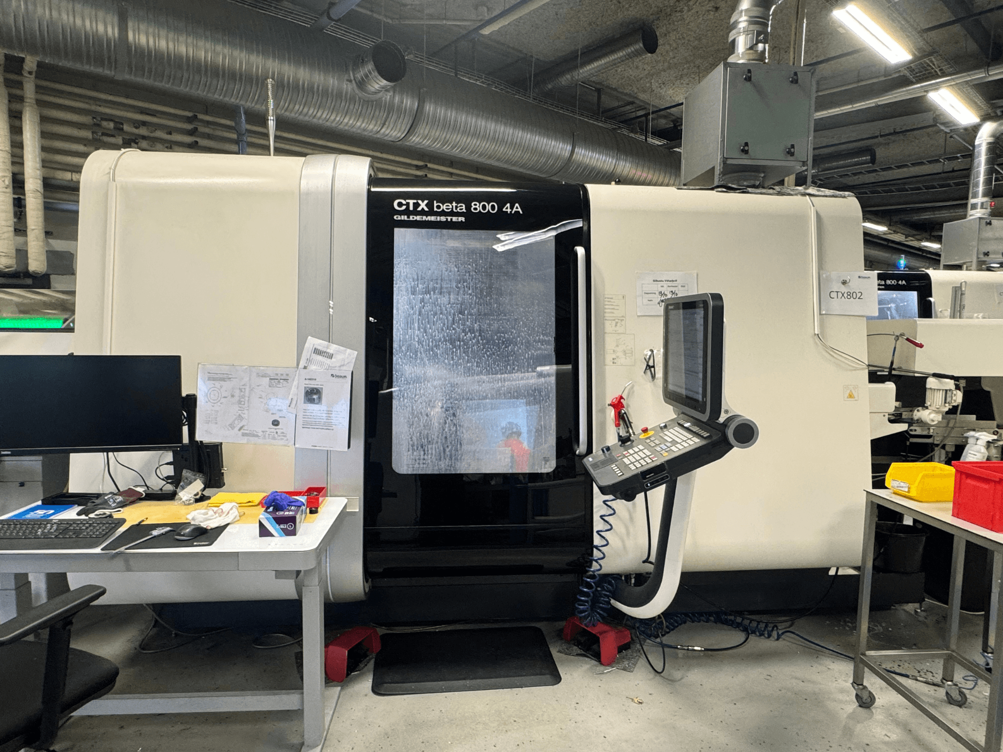 Maszyna CNC CTX beta 800 4A w warsztacie, widok z boku przedstawiający panel sterowania i przestrzeń roboczą zawaloną narzędziami i dokumentami.