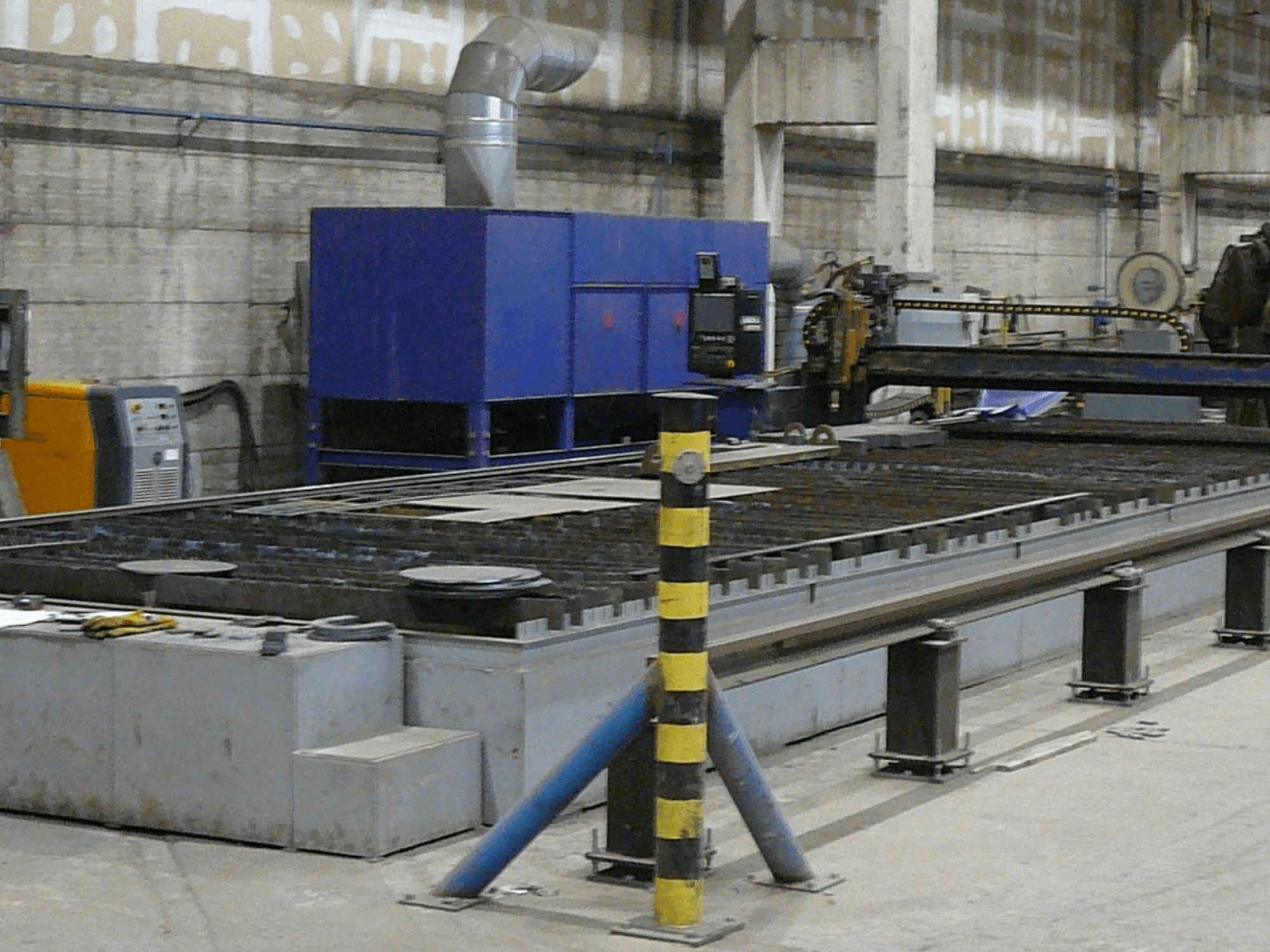 Przemysłowa maszyna do cięcia metalu, widok z boku, z niebieską skrzynką kontrolną, paskami bezpieczeństwa i metalową powierzchnią roboczą.
