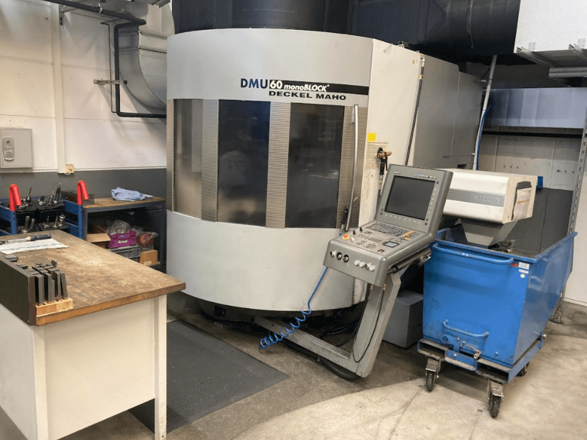 Pionowe centrum obróbcze DMG MORI DMU 60 Monoblock, widok z przodu z widocznym panelem sterowania, obszarem roboczym i niebieskim wózkiem narzędziowym.