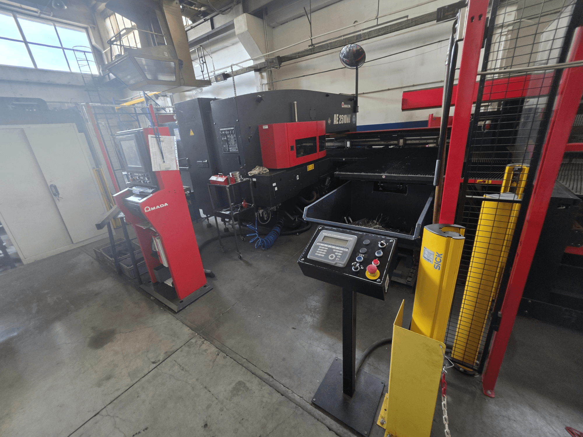 Wykrawarka CNC AMADA AE-2510NT – widok z przodu z panelem sterowania, elementami zabezpieczającymi oraz otaczającym ją otoczeniem przemysłowym.