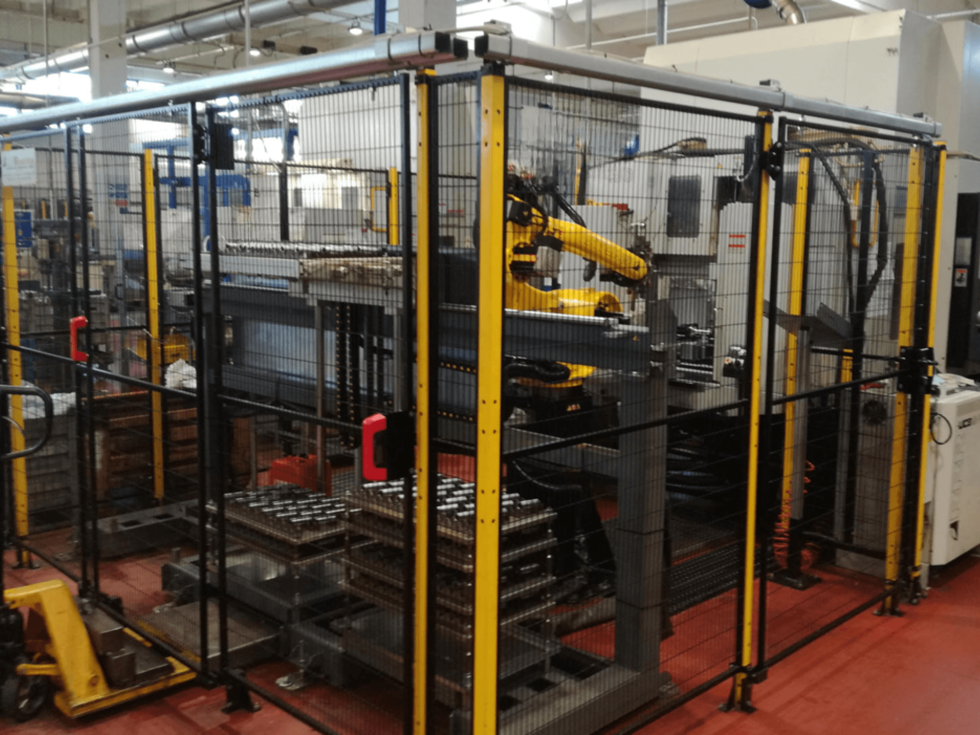 Pionowe centrum obróbcze Enshu EV450 w ogrodzonym obszarze roboczym, wyposażone w ramiona robotyczne i wiele ułożonych w stos przedmiotów obrabianych. Widok z przodu.
