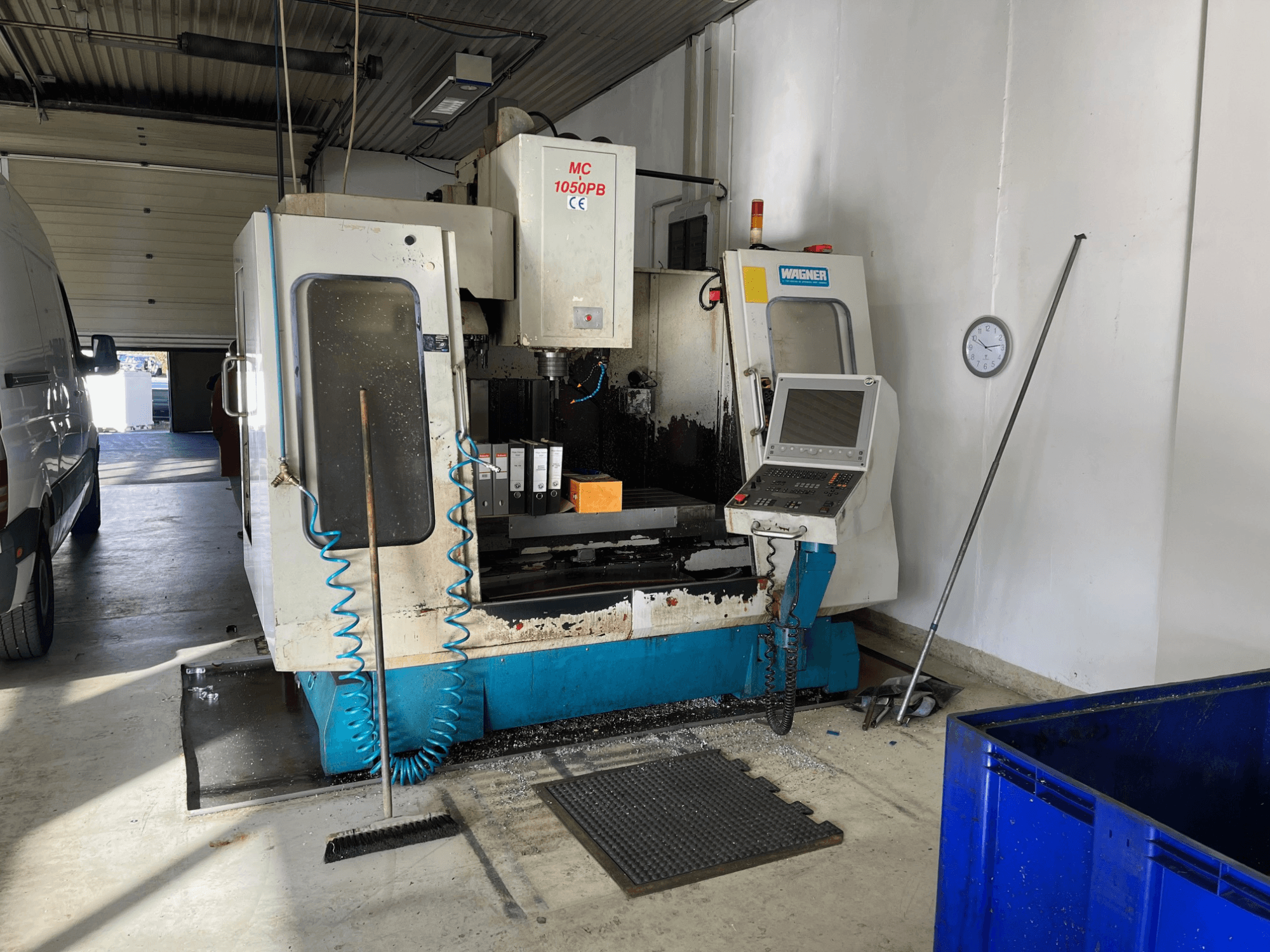 Maszyna CNC WAGNER MC 1050PB w warsztacie, widok z przodu, z panelem sterowania i obszarem roboczym; widoczny niebieski wąż powietrza i narzędzia czyszczące.