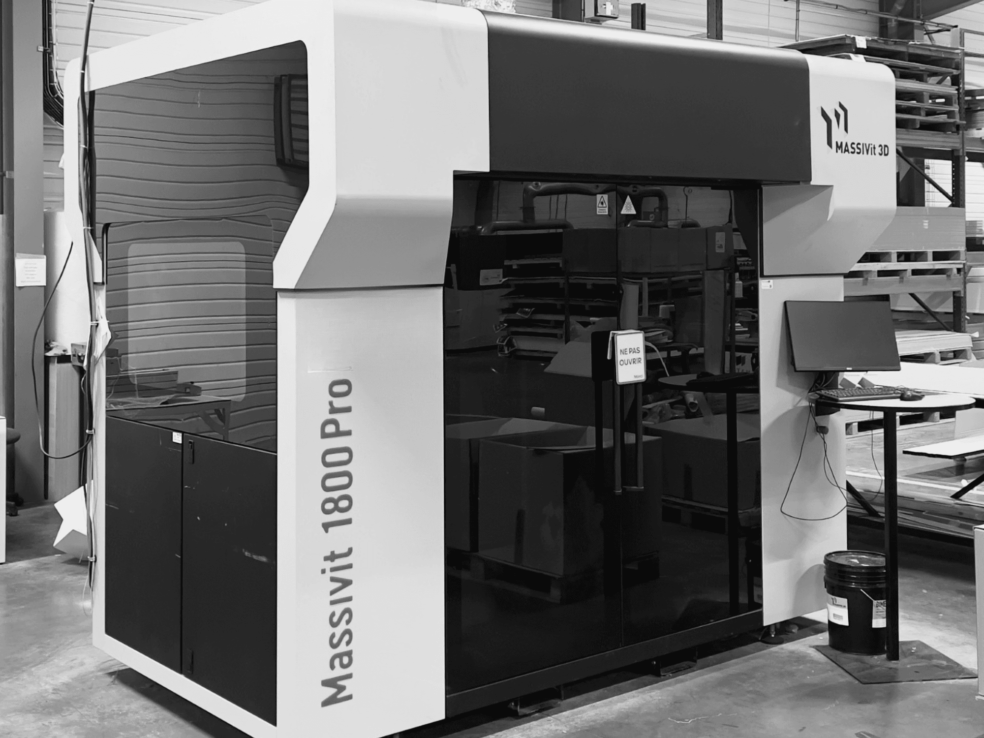 Drukarka 3D do tworzyw sztucznych MASSIVIT 1800 PRO w warsztacie, widok z przodu, z widoczną stacją roboczą z komputerem i panelem sterowania.