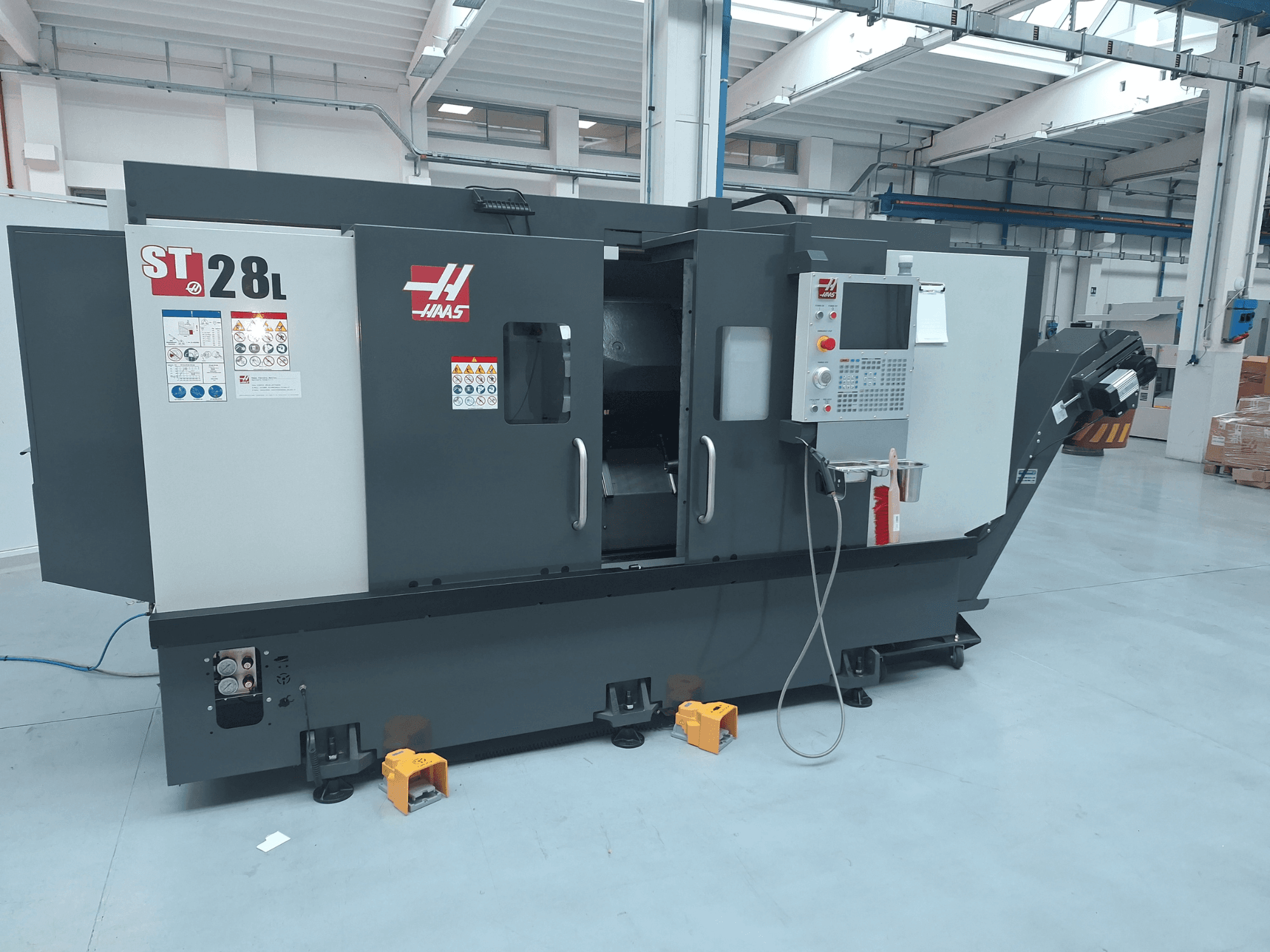 Widok z boku poziomej tokarki Haas ST-28L, przedstawiający panel sterowania, przyciski obsługowe oraz etykiety bezpieczeństwa.