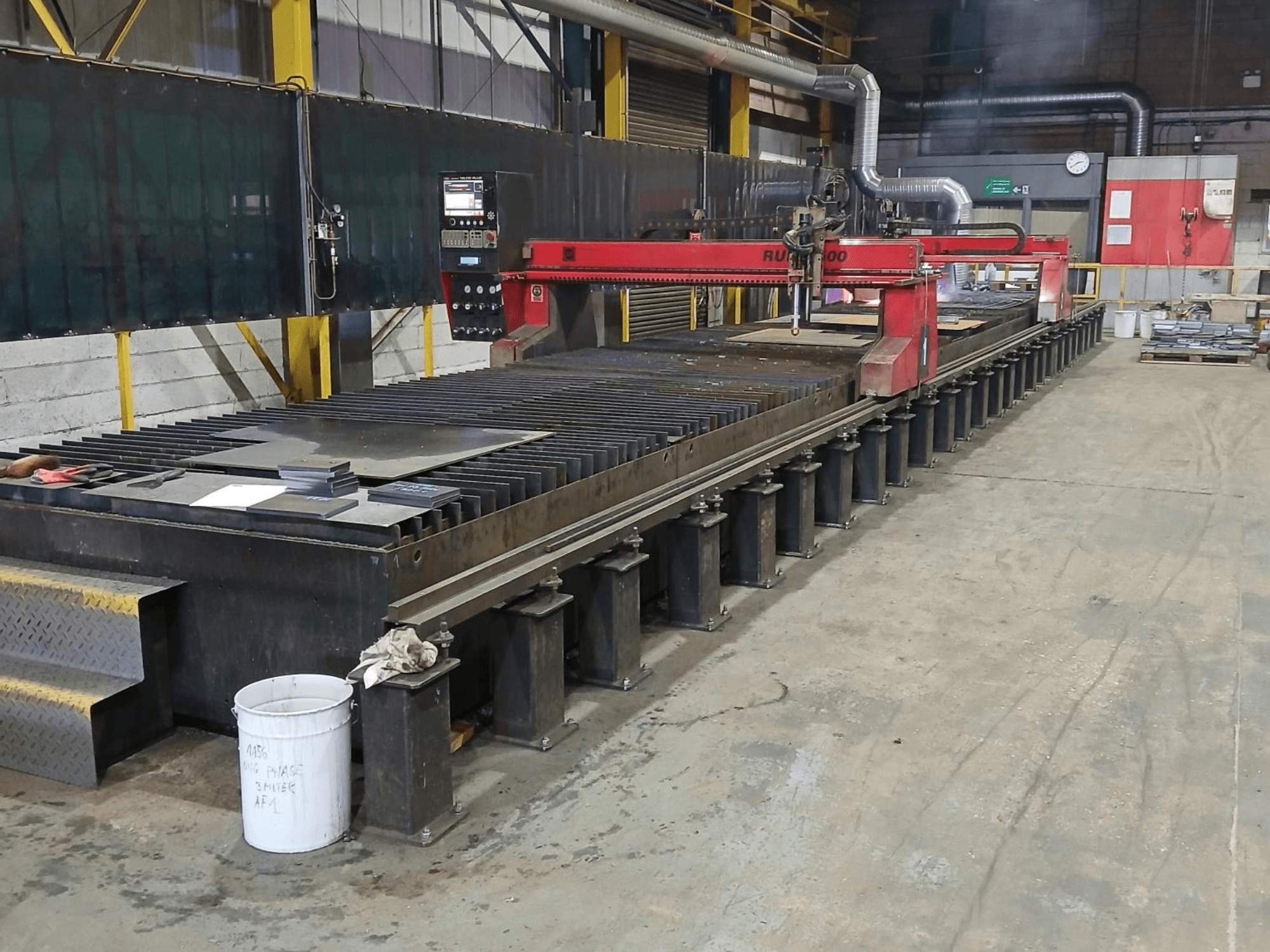 Czerwona przecinarka RUIDA 300, widok z boku, z blachą na stalowym stole, umieszczona w przemysłowym obszarze roboczym.