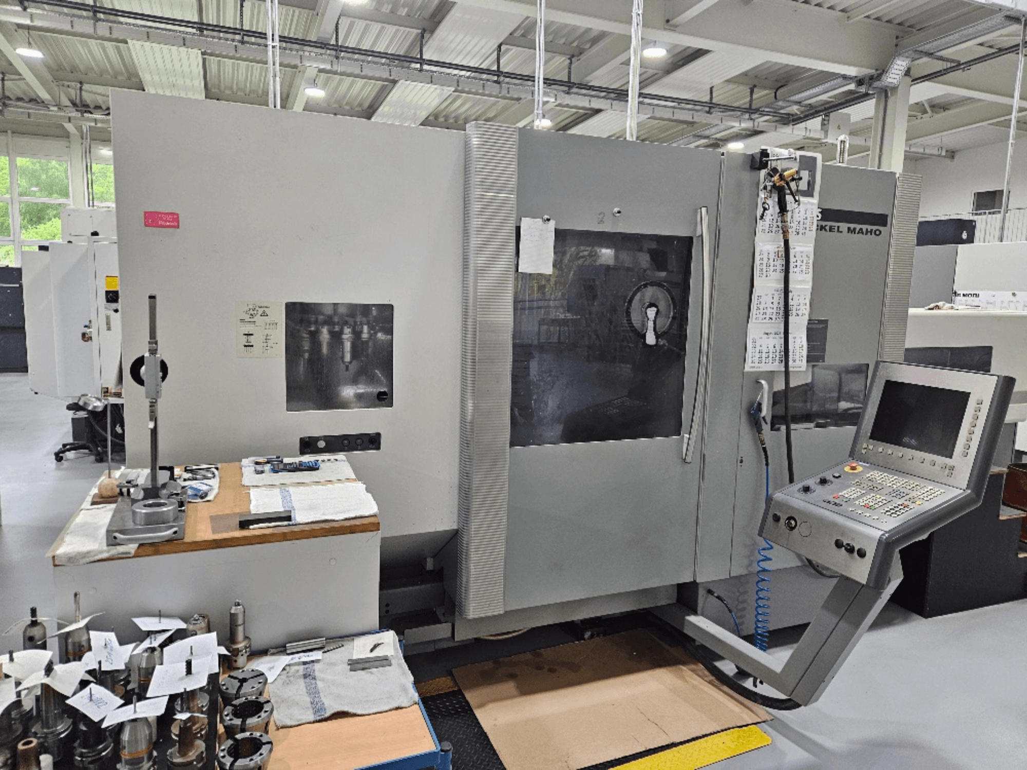 Widok z przodu maszyny DMG Mori DMU 70 z panelem sterowania, narzędziami na drewnianym stole i różnymi przedmiotami obrabianymi w pobliżu.