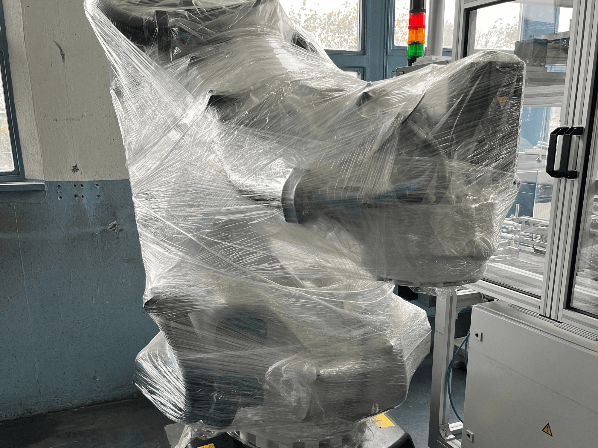 Przemysłowe ramię robota owinięte tworzywem sztucznym, widok z przodu. Zawiera jednostkę sterującą i lampkę kontrolną. Marka niewidoczna.