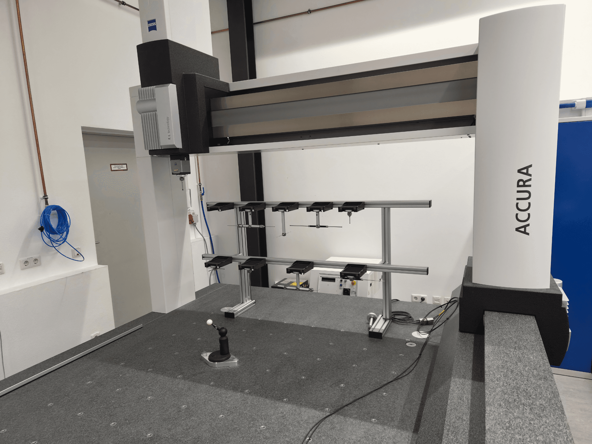 Maszyna CMM ZEISS ACCURA 16/24/10 widziana z przodu, z widocznym ramieniem pomiarowym, podstawą i panelem sterowania na szarej powierzchni pokrytej wykładziną.