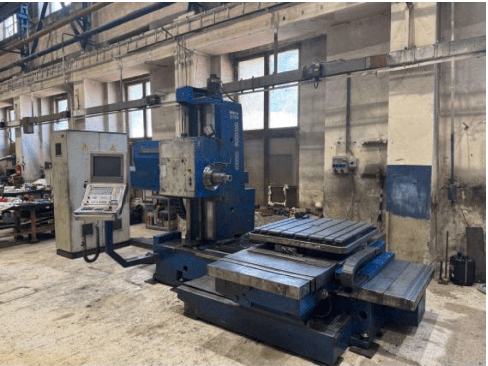 Widok z przodu wytaczarki CNC TOS WHN 9 B, wyposażonej w panel sterowania i duży stół roboczy, umieszczonej w środowisku warsztatowym.