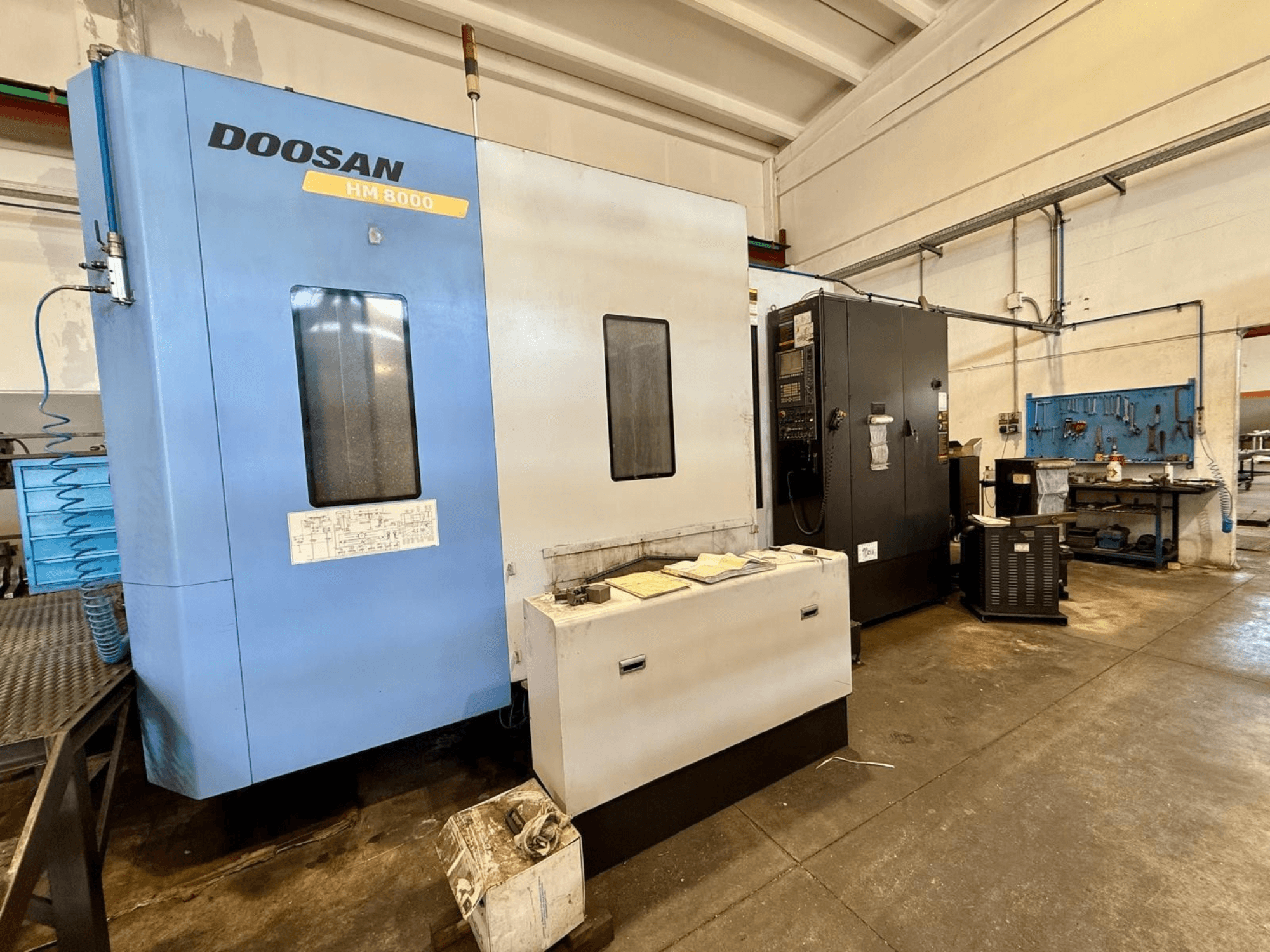 Poziome centrum obróbcze DOOSAN HM 8000 w kolorze niebieskim, widok z przodu, przedstawiający panel sterowania i znajdujące się w pobliżu urządzenia warsztatowe.