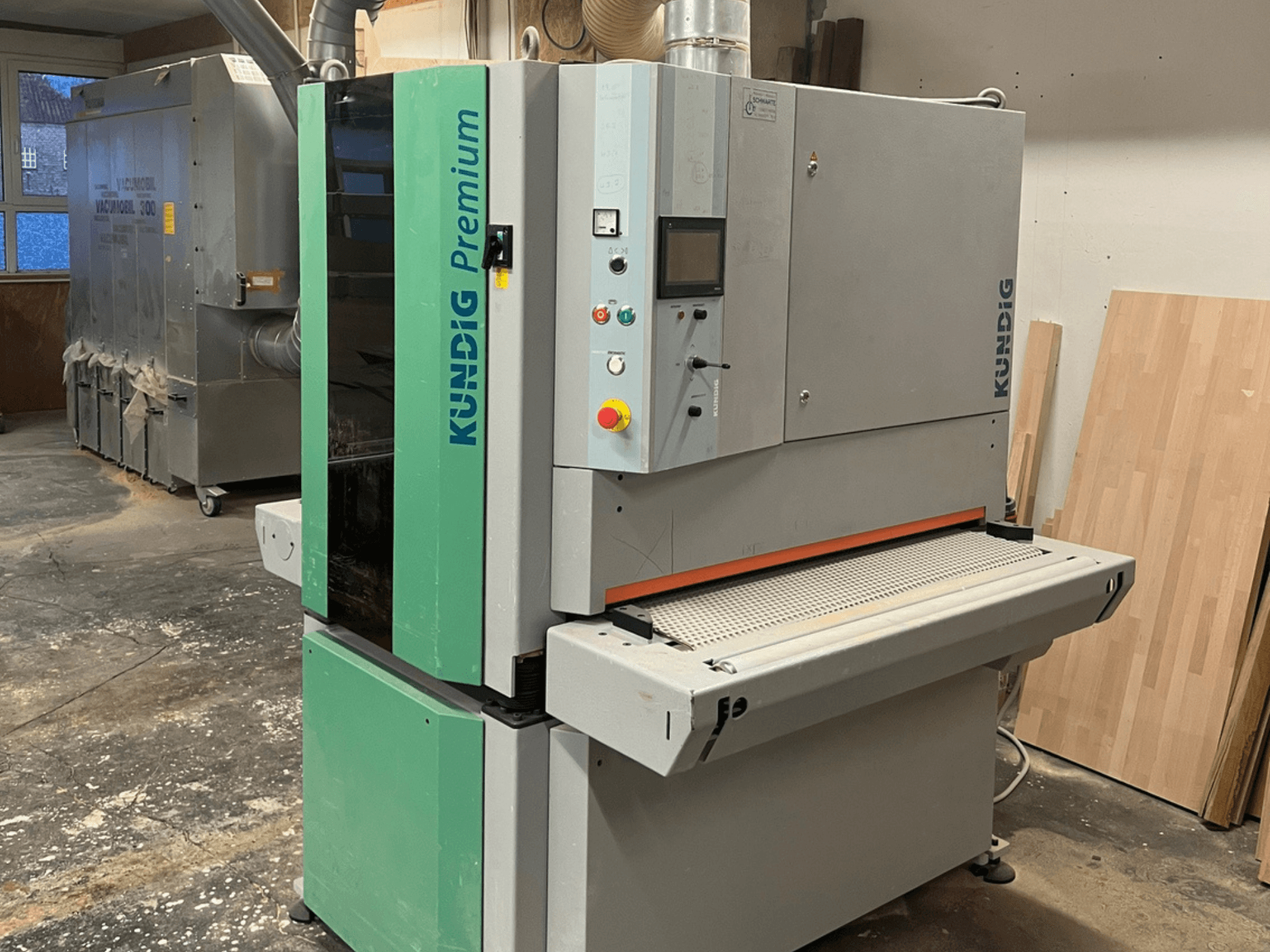 Szlifierka taśmowa Kundig Premium-1 1100K w kolorze szaro-zielonym, pokazana z boku z widocznym panelem sterowania i taśmą szlifierską.