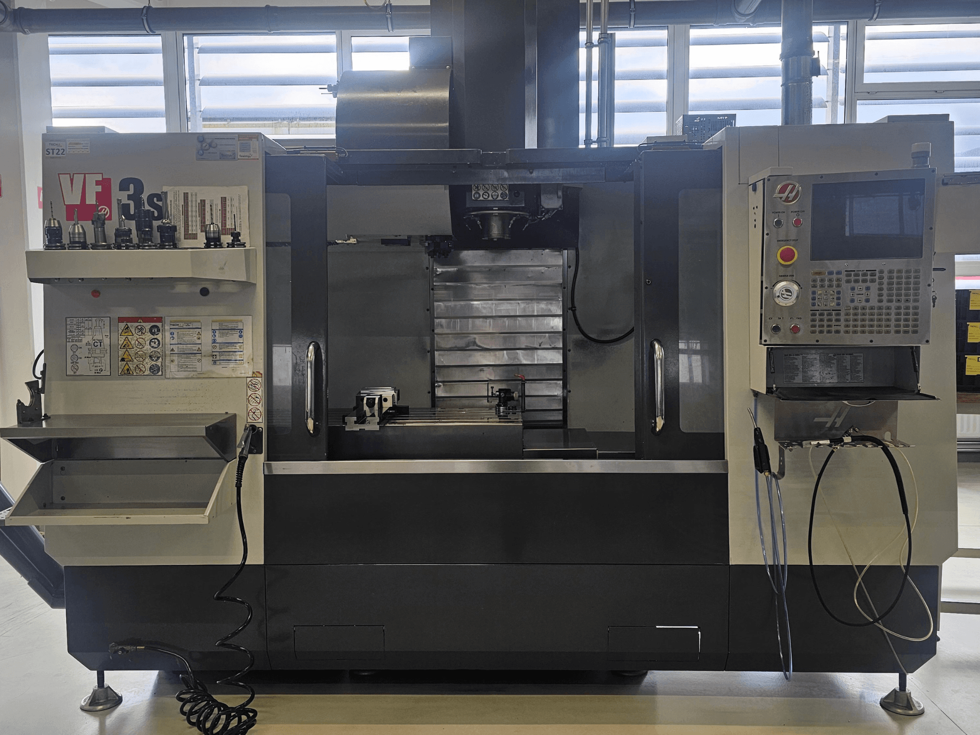 Frezarka CNC Haas VF3SS, widok z przodu, z uchwytami narzędzi, panelem sterowania i pustą tacą na chłodziwo, z widocznymi etykietami bezpieczeństwa.