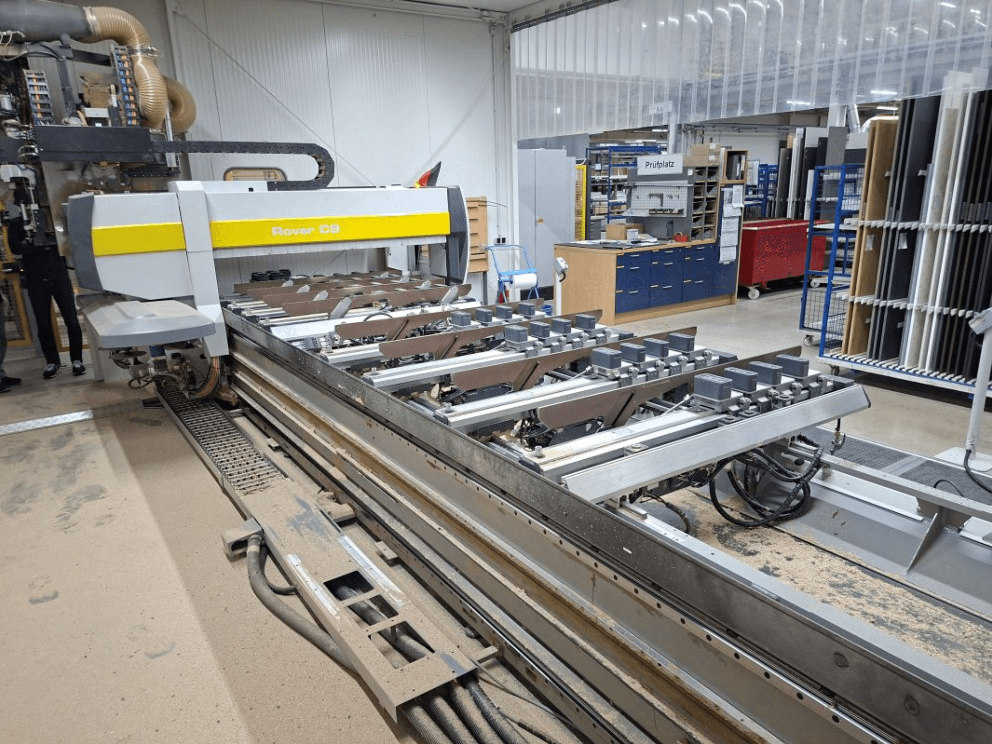 Maszyna CNC Rover C9 w warsztacie; widok z przodu przedstawia narzędzia tnące i system przenośników, z pyłem gromadzącym się na podłodze.