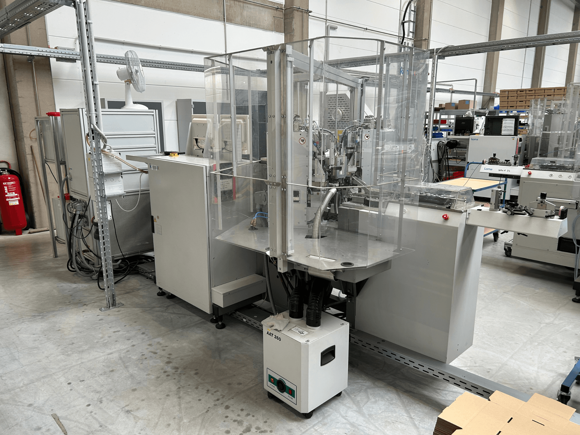 Urządzenie automatyzacyjne KOMAX Alpha 433 L w warunkach fabrycznych. Wyposażone jest w przezroczystą obudowę zabezpieczającą oraz różne panele sterowania.