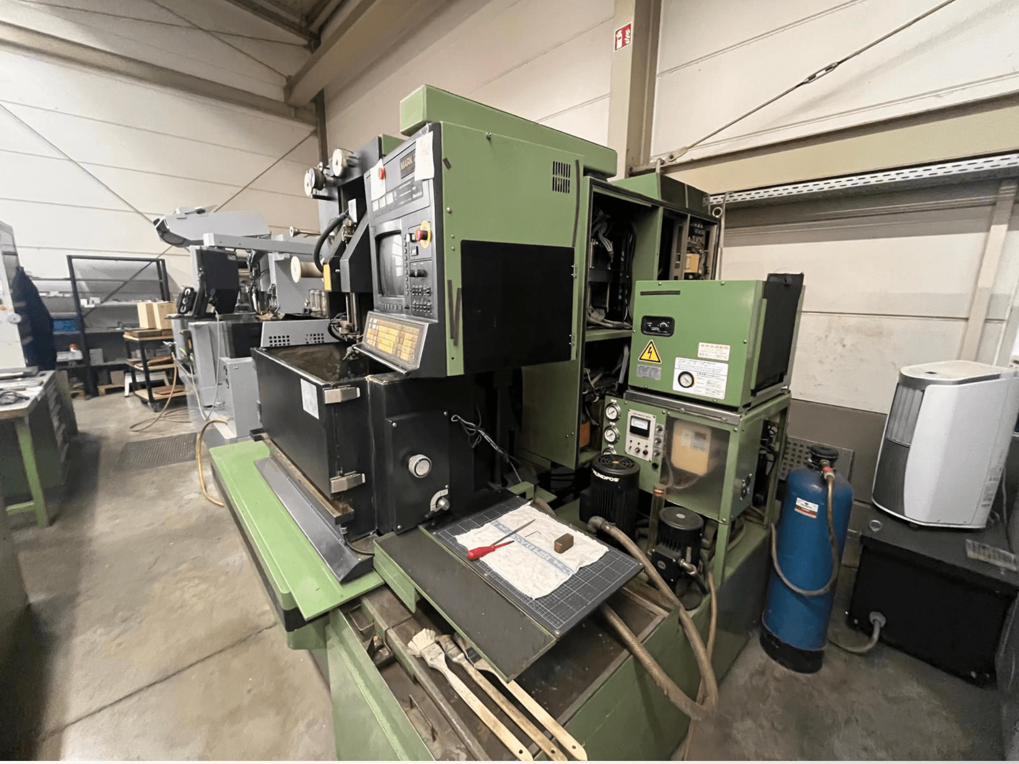 Zielona maszyna przemysłowa z panelem sterowania, widok z boku; zawiera elementy hydrauliczne i powierzchnię roboczą z narzędziami.