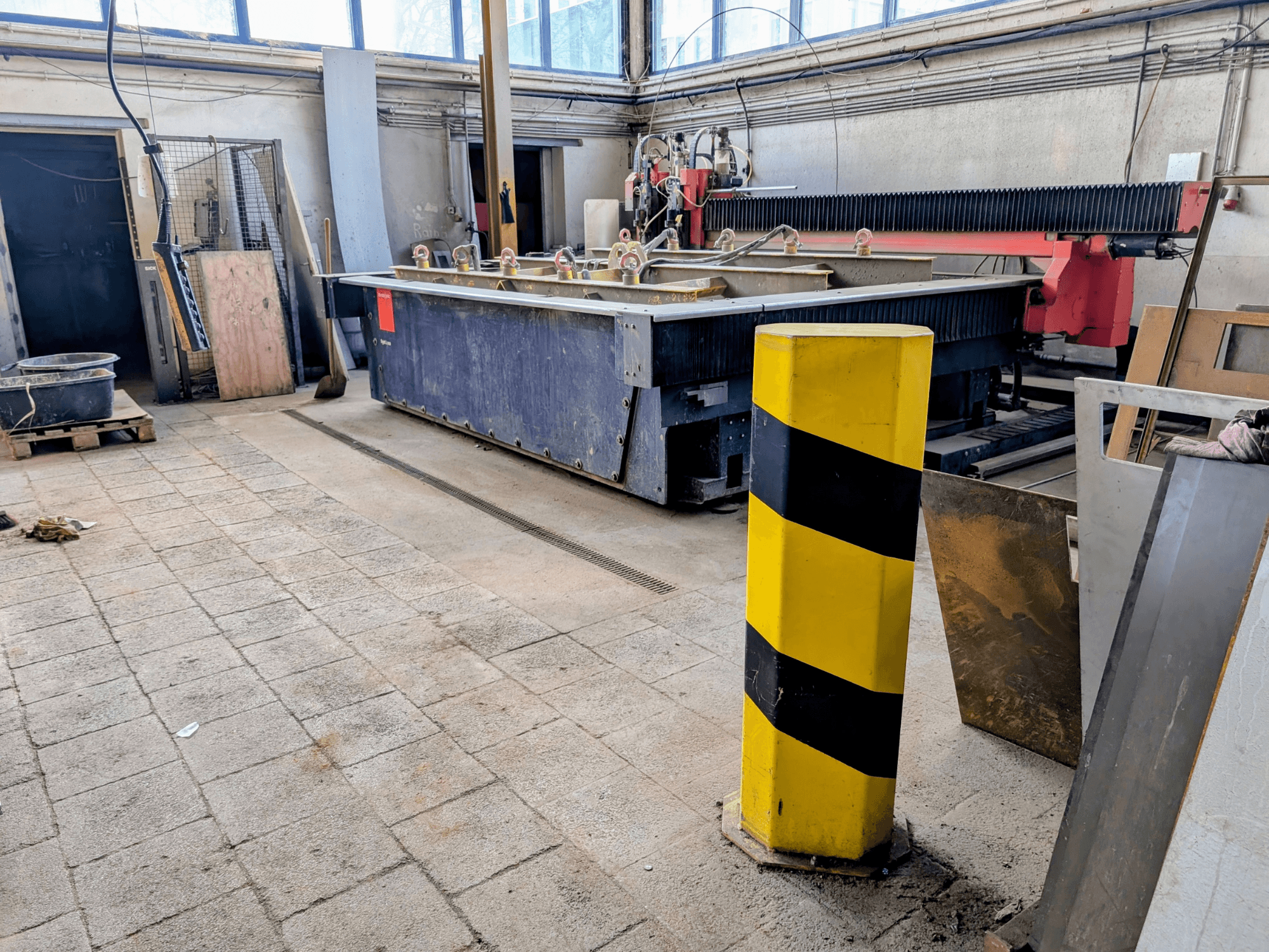 Maszyna do cięcia strumieniem wody Bystronic Byjet L 2030 w warsztacie, widok z boku, z żółtym słupkiem bezpieczeństwa i otaczającymi materiałami.