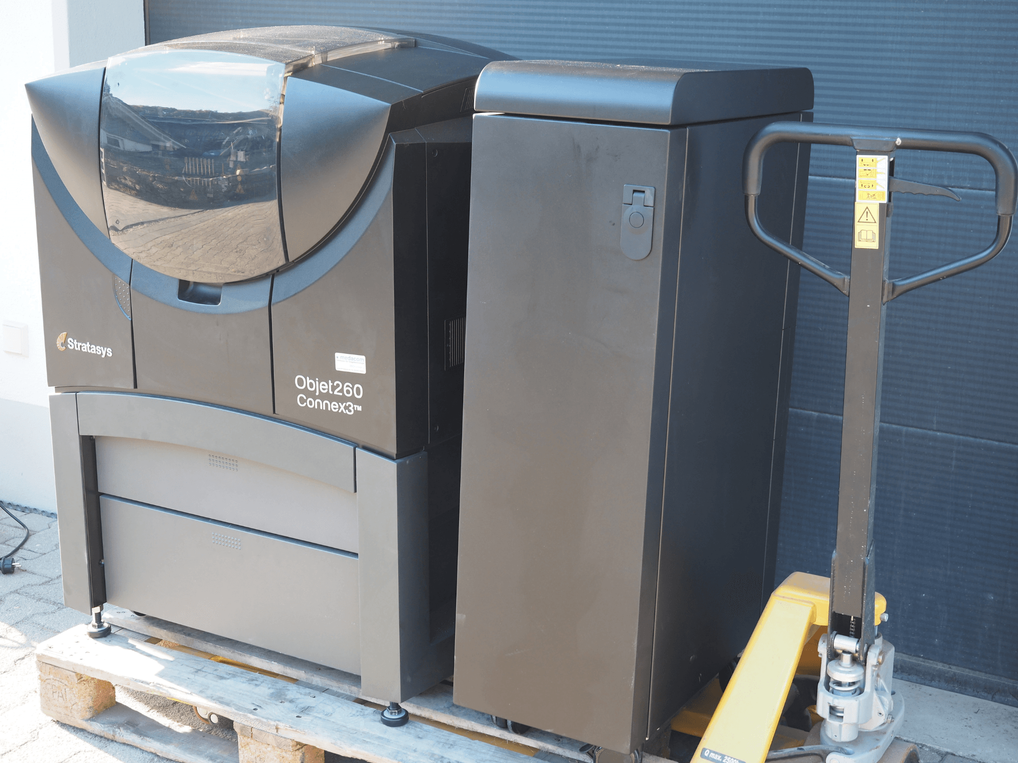 Drukarka 3D do tworzyw sztucznych Stratasys Objet260 Connex3, widok z boku, charakteryzuje się kompaktową konstrukcją z panelem sterowania i podłączonym pojemnikiem na materiał.