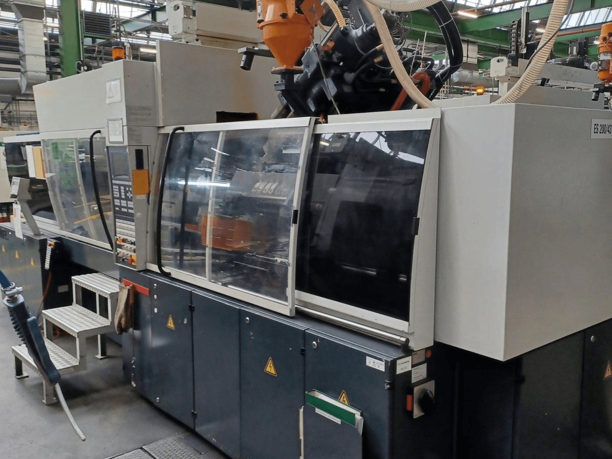 Widok z przodu wtryskarki do tworzyw sztucznych, marka i model nieznane, z przezroczystymi drzwiami bezpieczeństwa i panelem sterowania.