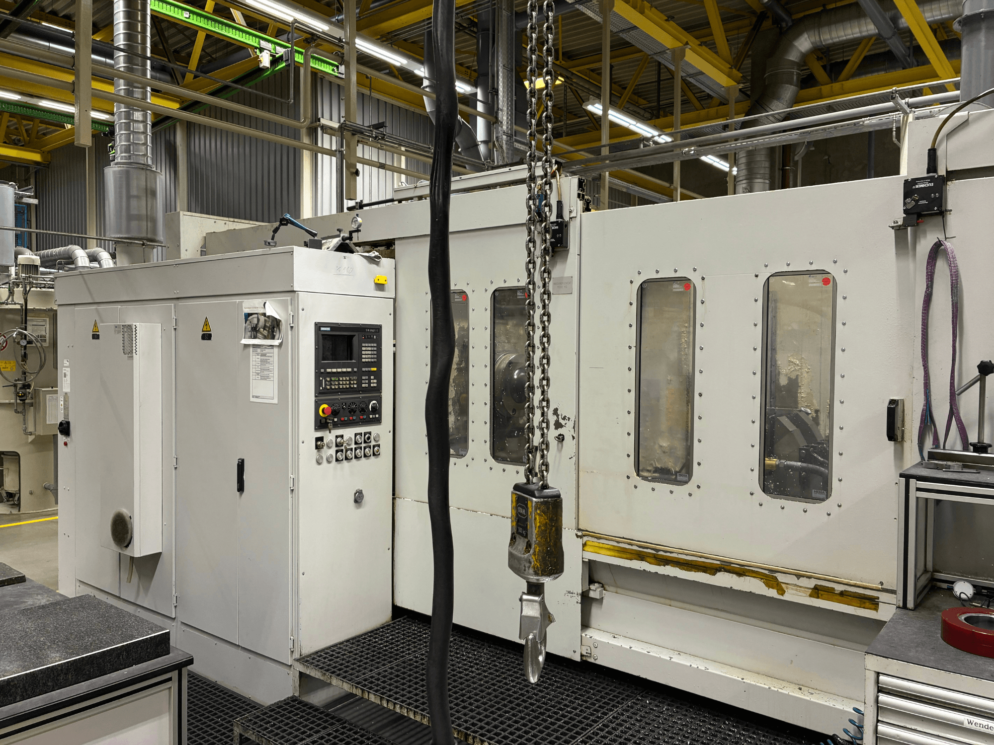 Przemysłowa maszyna CNC z panelem sterowania, widok z boku; zawiera okna, łańcuchy i elementy metalowe w otoczeniu fabrycznym.